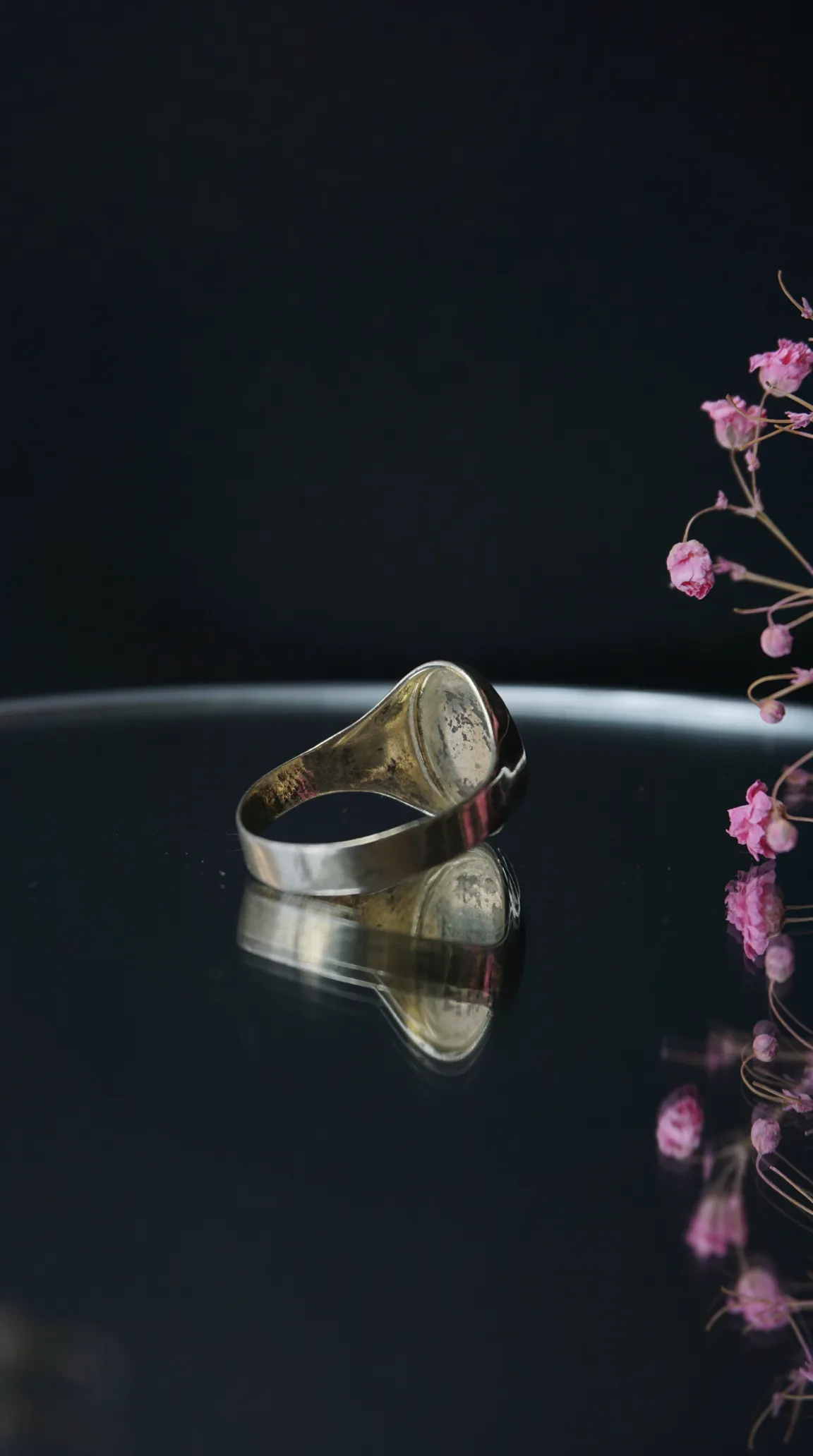 Vintage silver-tone signet ring with aged patina on a dark reflective surface, with dried pink flowers.