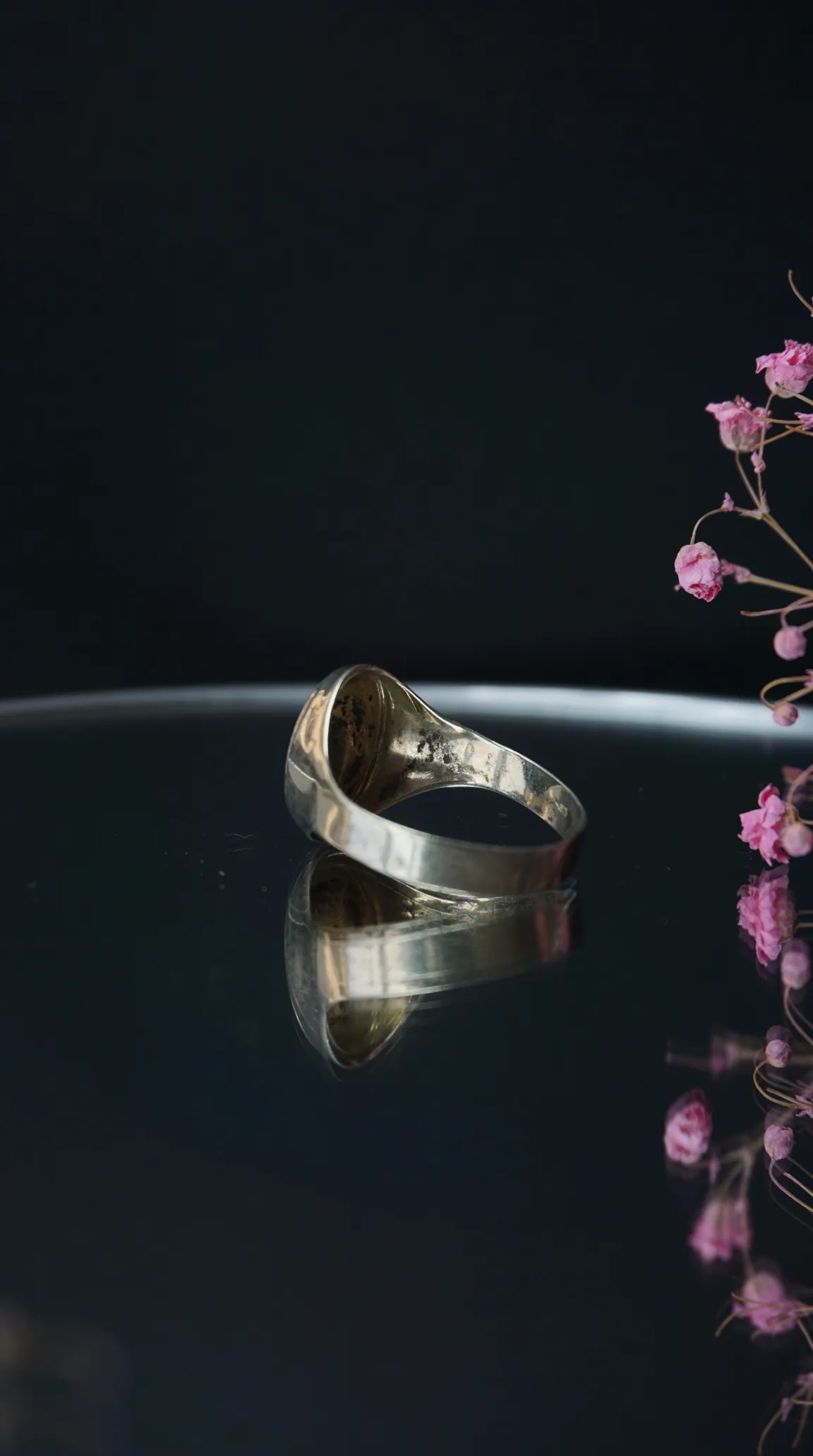 Vintage silver ring with reflection on dark surface, accented by dried pink flowers.