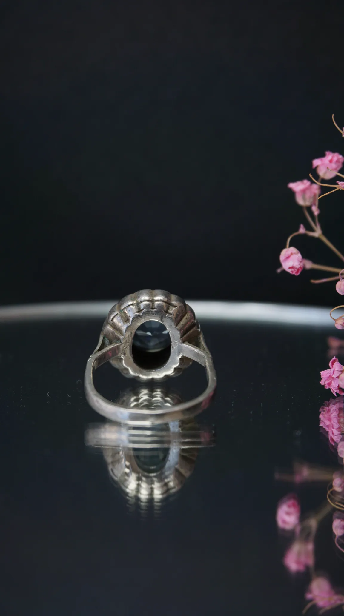 Vintage silver ring with dark blue oval gemstone and scalloped setting, on a reflective black background with pink dried flowers.