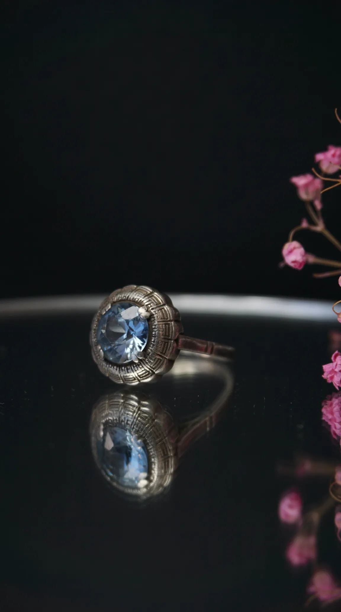 Vintage silver ring with light blue gemstone, intricate bezel design, reflecting on dark surface with pink flowers.