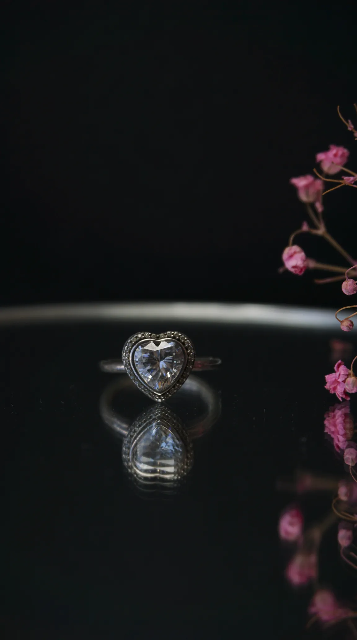 Vintage silver ring with heart-shaped clear gemstone and marcasite halo, reflecting on a black surface with pink floral accents.