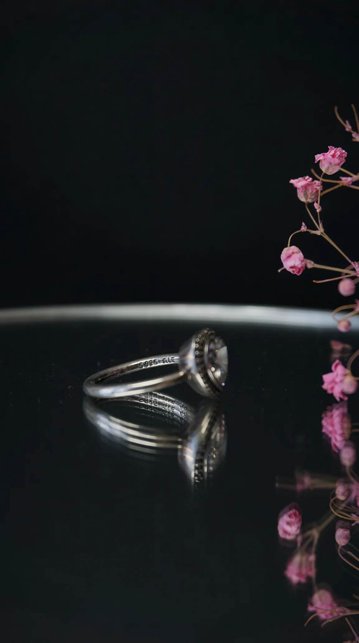 Vintage Pandora sterling silver ring with clear halo gemstone, reflected on a dark surface, beside delicate pink flowers.