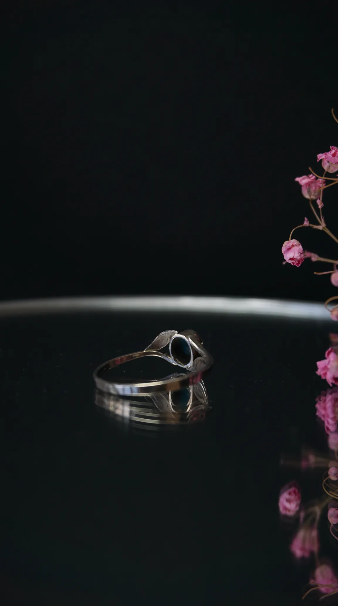 Vintage silver ring with oval dark gemstone and delicate leaf details, resting on a reflective dark surface with pink flowers.