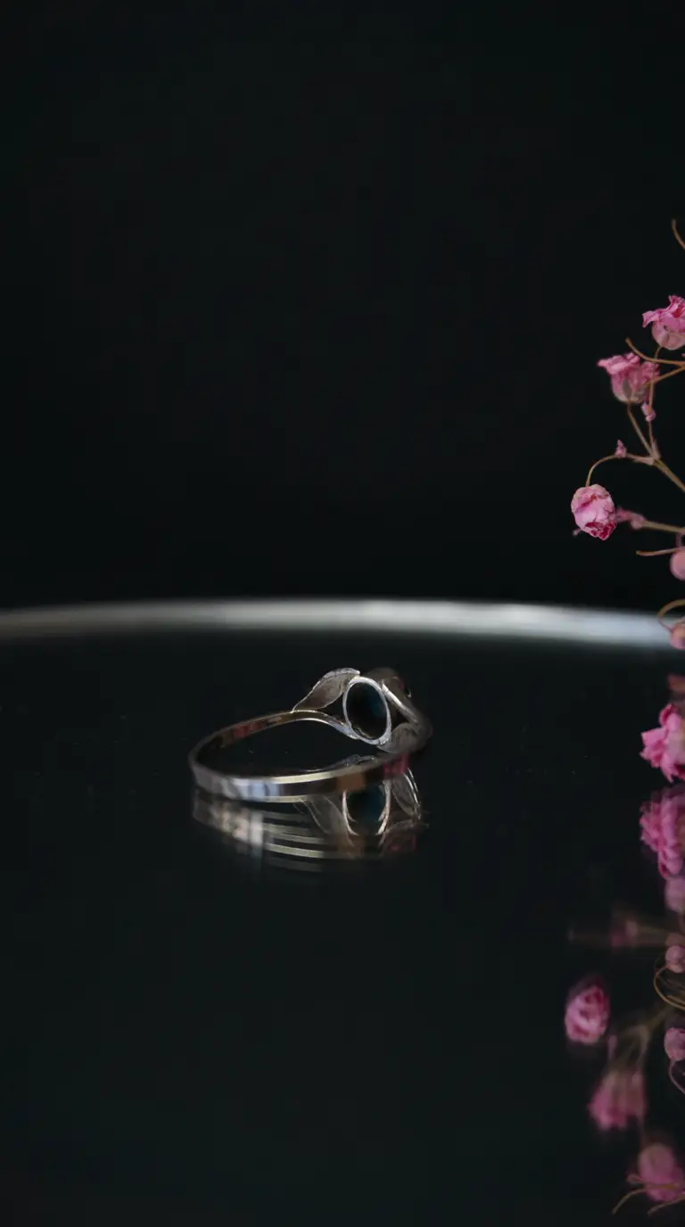 Vintage silver ring with oval dark gemstone and delicate leaf details, resting on a reflective dark surface with pink flowers.