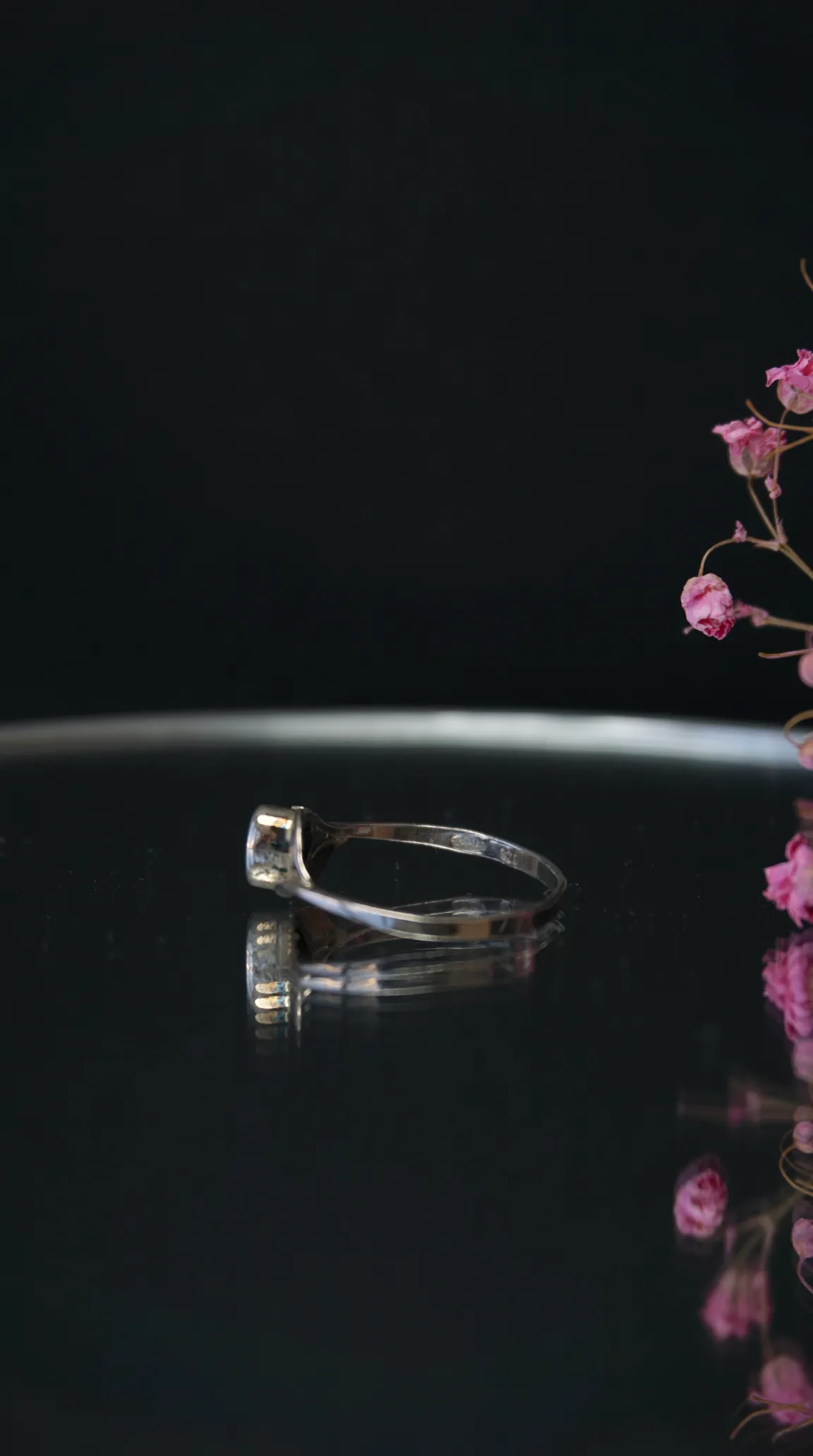 Vintage silver ring with a small setting, reflecting on a dark surface, next to dried pink flowers.
