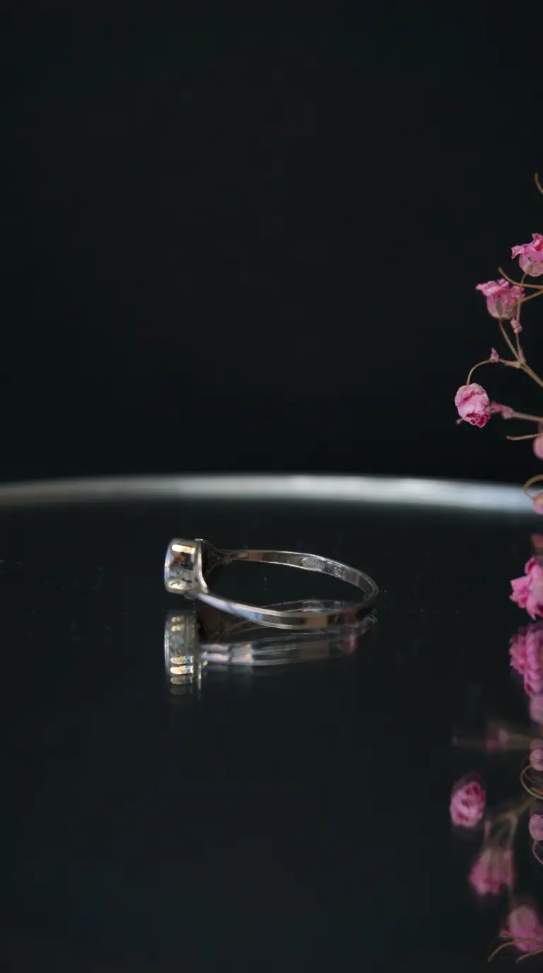 Vintage silver ring with a small setting, reflecting on a dark surface, next to dried pink flowers.