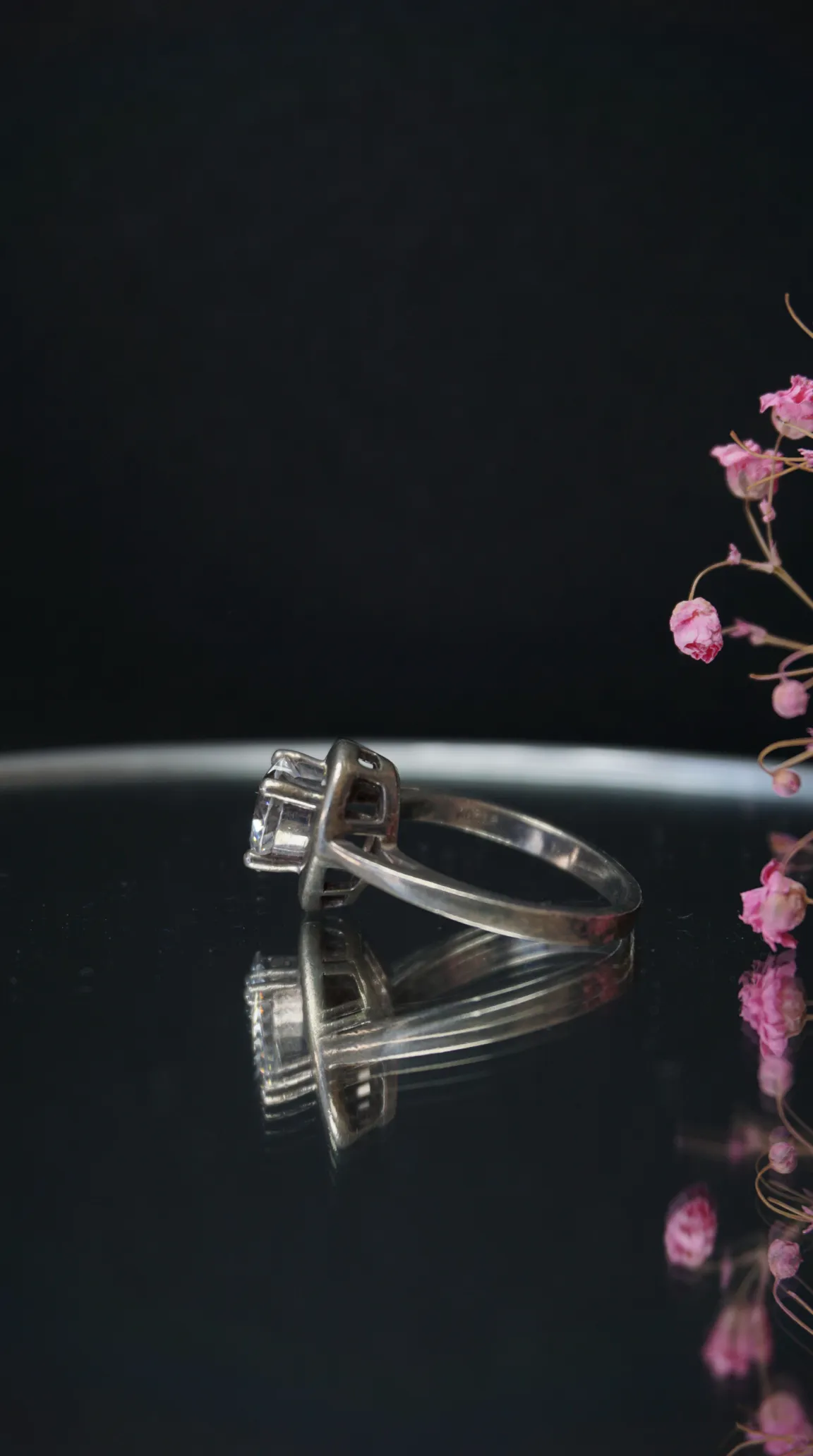 Vintage silver solitaire ring with clear stone on reflective surface, pink floral accents.