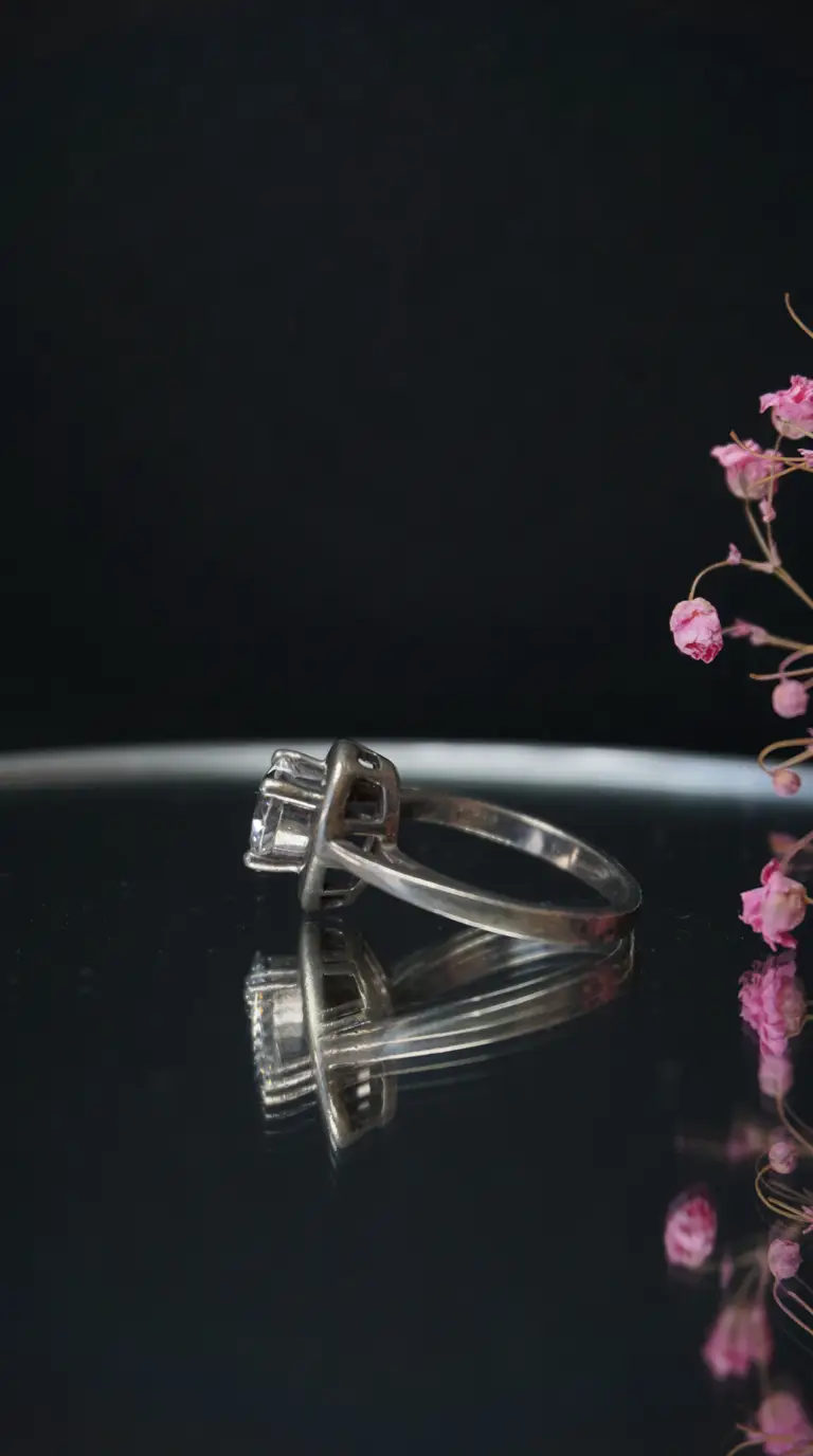 Vintage silver solitaire ring with clear stone on reflective surface, pink floral accents.