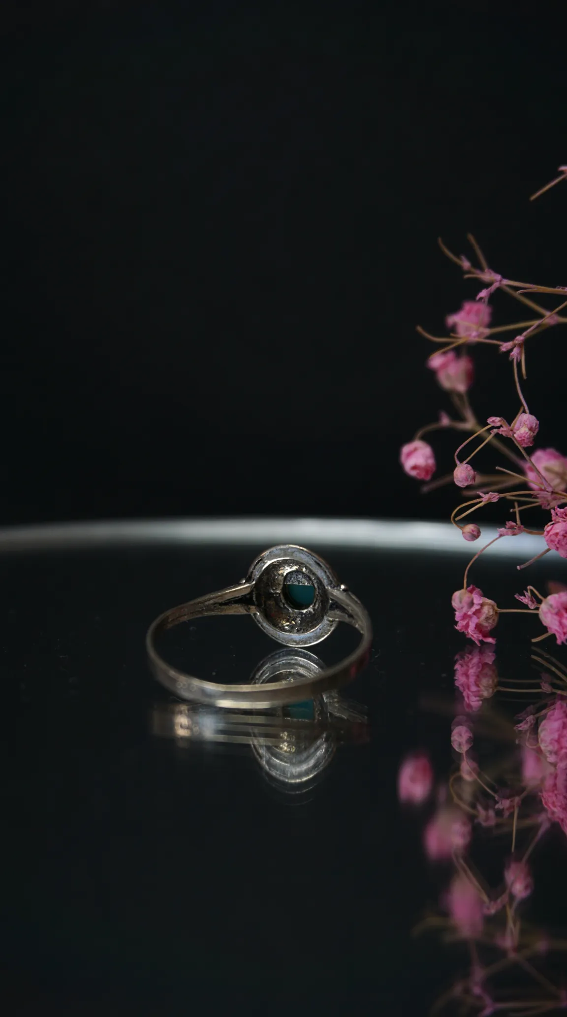 Vintage sterling silver ring with a round teal gemstone, displayed on a reflective black surface with dried pink flowers.