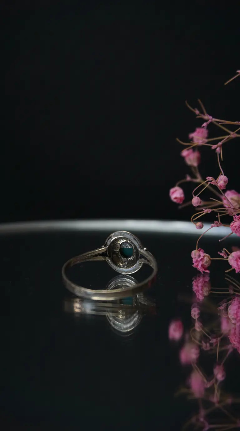 Vintage sterling silver ring with a round teal gemstone, displayed on a reflective black surface with dried pink flowers.