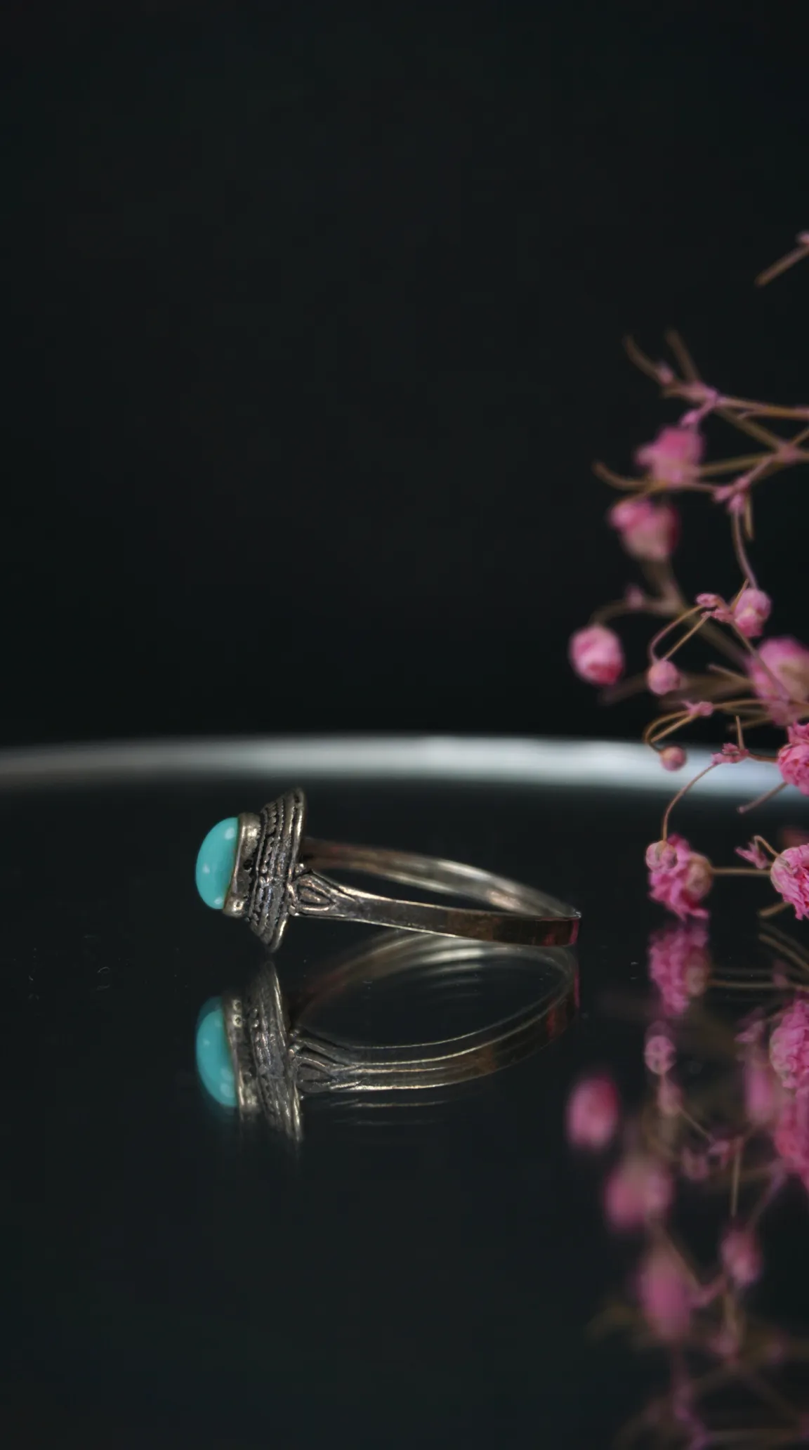 Vintage sterling silver ring with a round turquoise gemstone on a reflective dark surface, with soft pink dried flowers in the background.