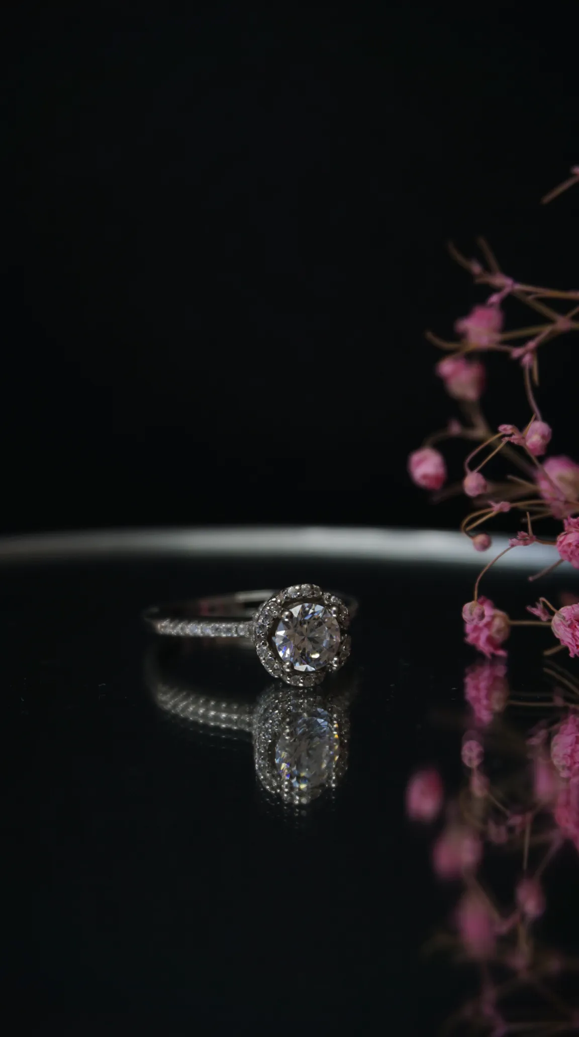 Vintage sterling silver halo ring with brilliant round clear gemstone and pave band, reflecting on a dark surface with delicate pink flowers.