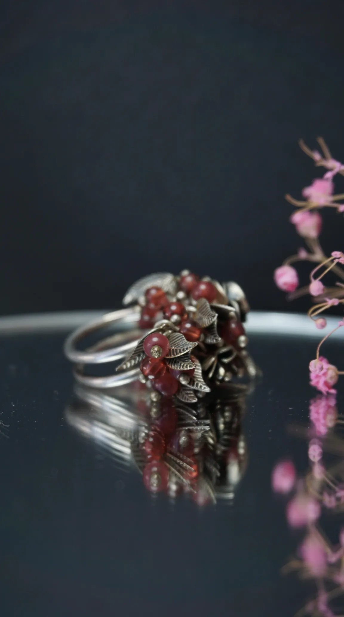 Vintage silver ring with red beaded cluster and delicate silver leaf accents, reflected on a dark polished surface, with soft pink floral details.