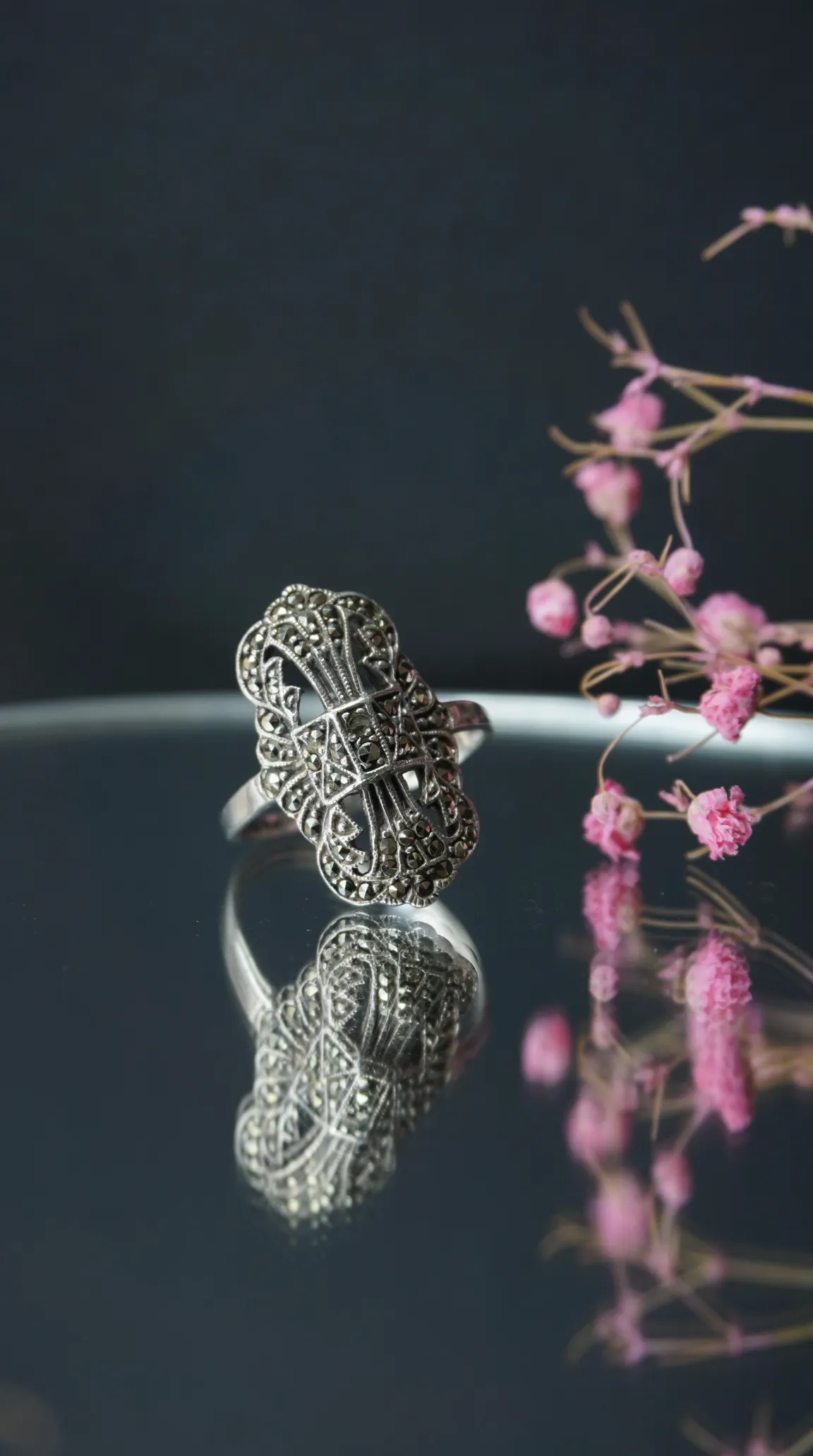 Vintage Art Deco sterling silver marcasite filigree ring reflecting on dark surface with pink flowers.
