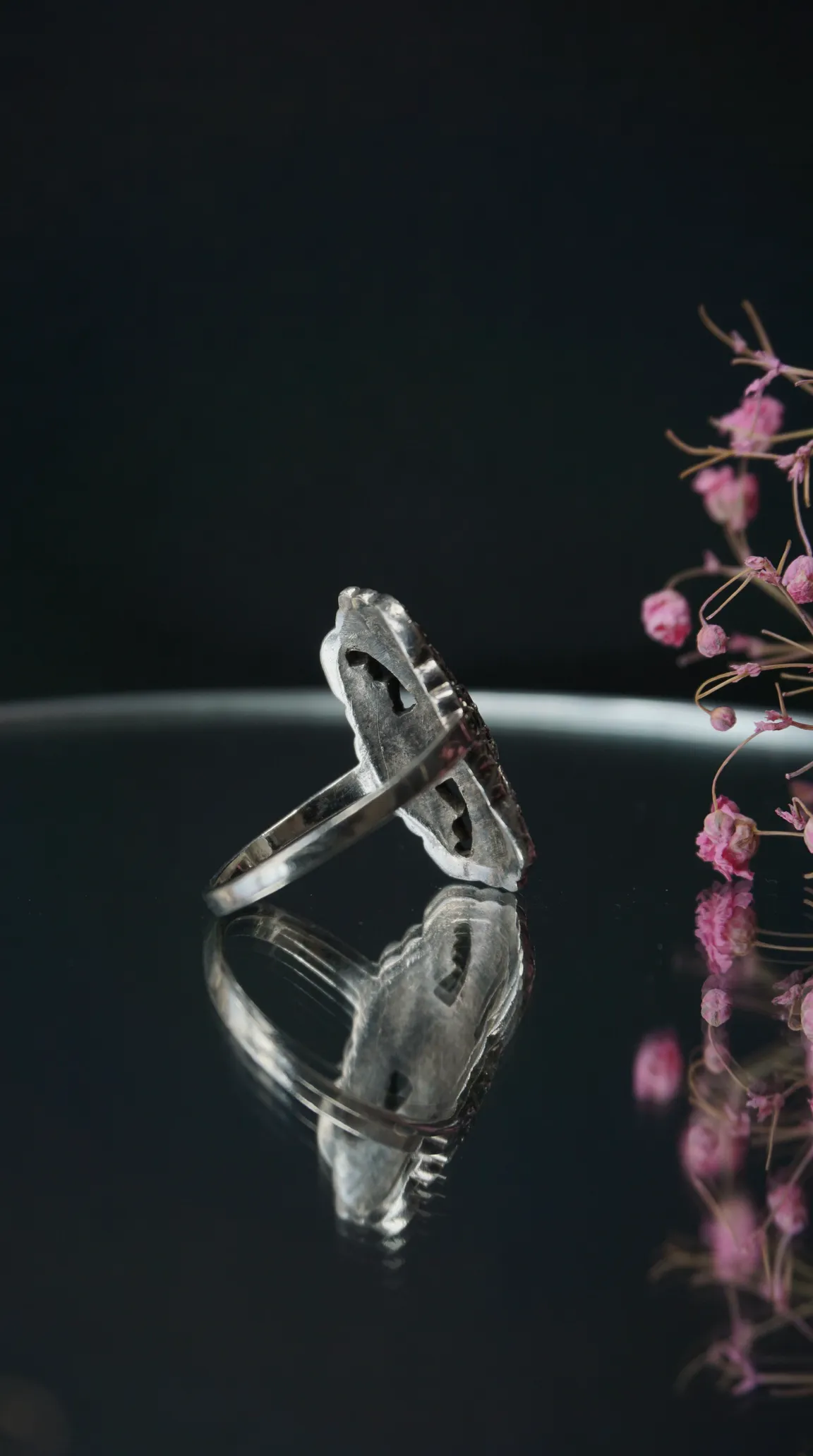 Vintage sterling silver ring with intricate design, side view, reflecting on a dark surface with pink dried flowers.