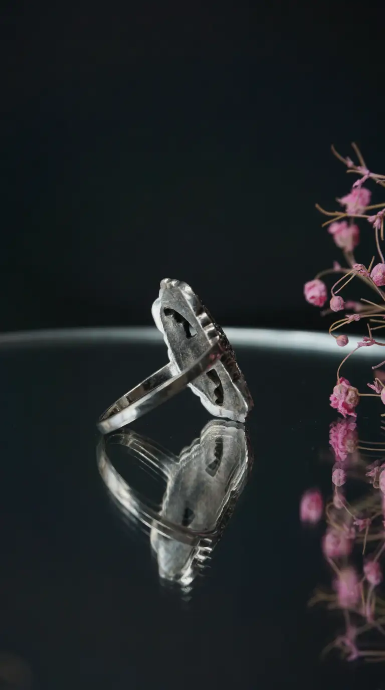 Vintage sterling silver ring with intricate design, side view, reflecting on a dark surface with pink dried flowers.