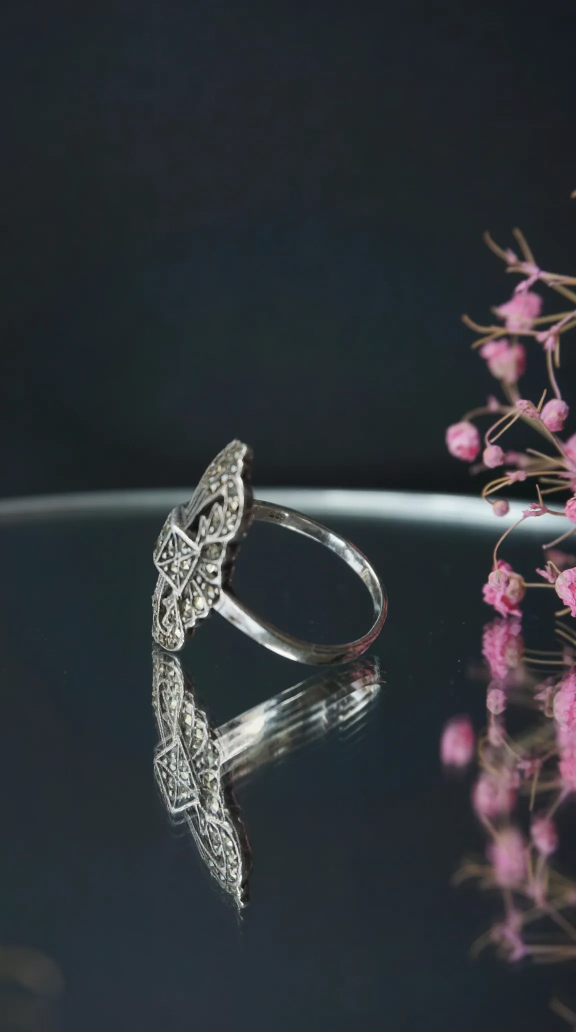 Vintage Art Deco silver marcasite ring with intricate geometric filigree, displayed on a reflective surface with soft pink floral accent.