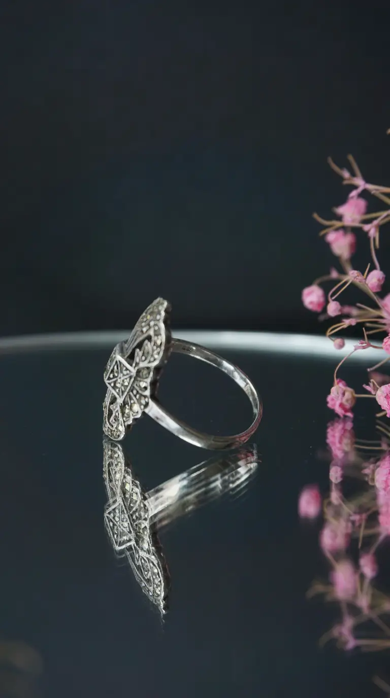Vintage Art Deco silver marcasite ring with intricate geometric filigree, displayed on a reflective surface with soft pink floral accent.