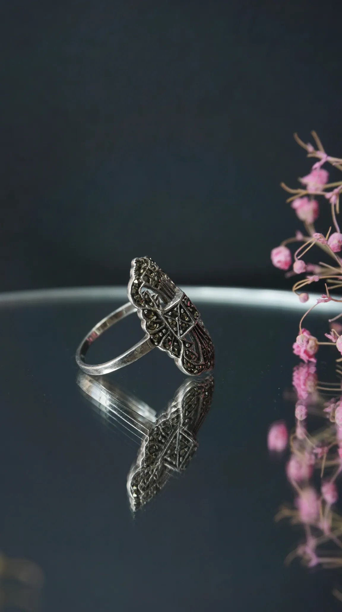 Vintage Art Deco marcasite silver ring with ornate design, reflected on a dark surface with pink floral accents.