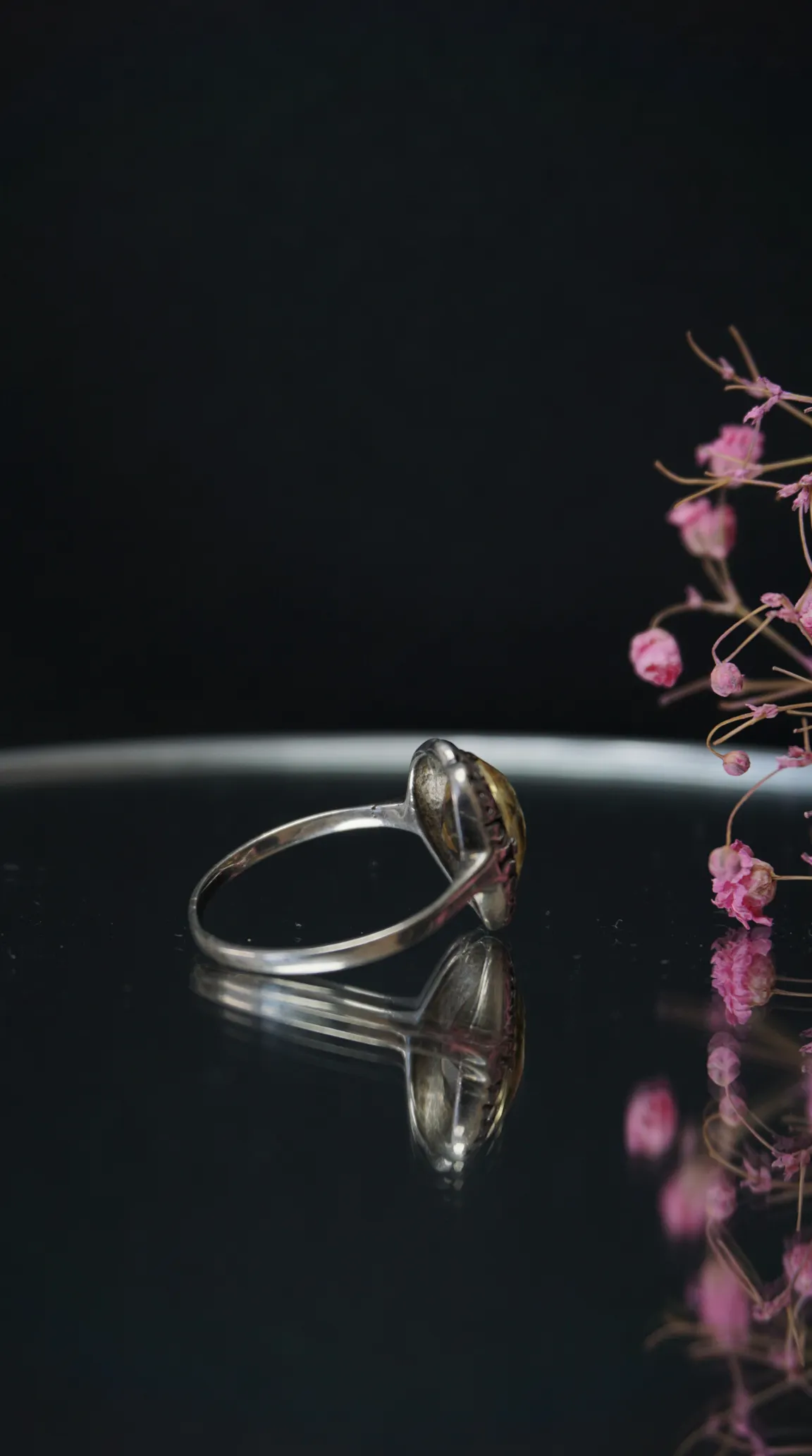 Vintage silver ring with golden heart motif on reflective black surface, accented by pink dried flowers