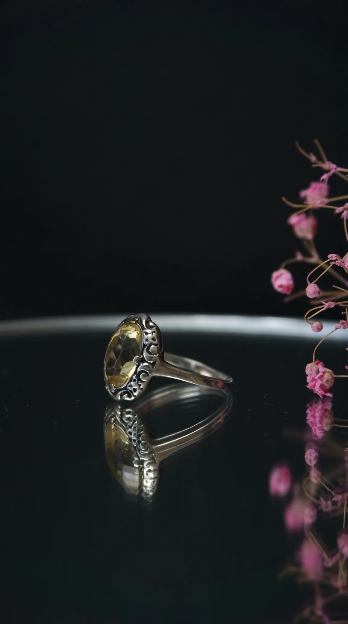 Vintage sterling silver ring with oval yellow citrine gemstone and filigree detail, reflected on dark surface with pink dried flowers.