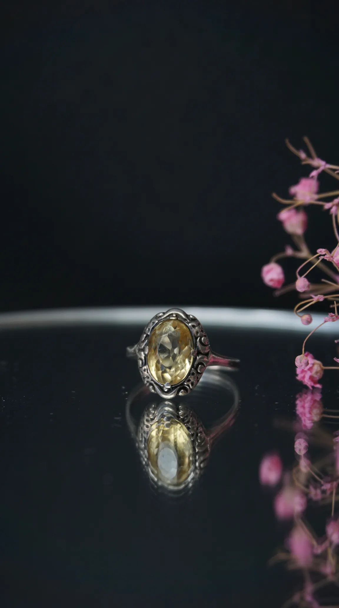 Vintage sterling silver ring with oval yellow citrine gemstone, ornate filigree setting, reflected on dark surface with pink flowers.