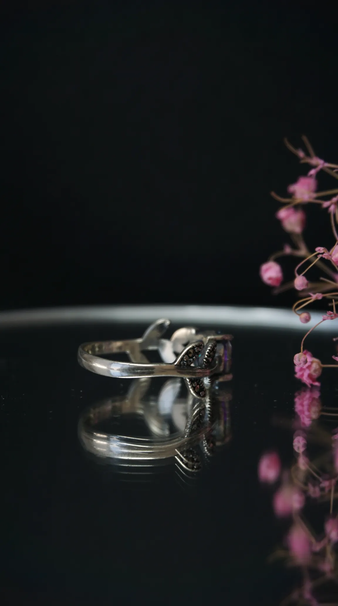 Vintage silver ring with iridescent gemstone and black marcasite accents, on a reflective surface, soft pink flowers in the background.