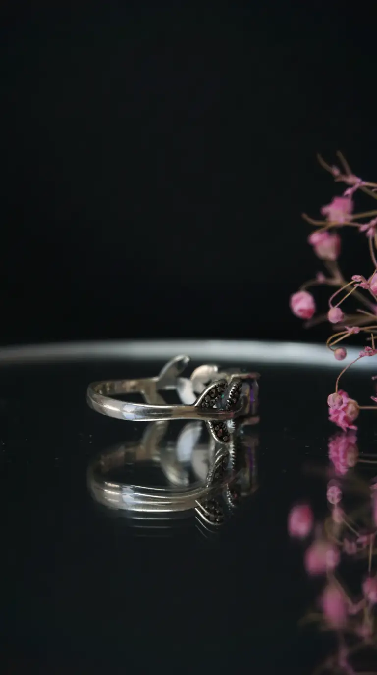 Vintage silver ring with iridescent gemstone and black marcasite accents, on a reflective surface, soft pink flowers in the background.