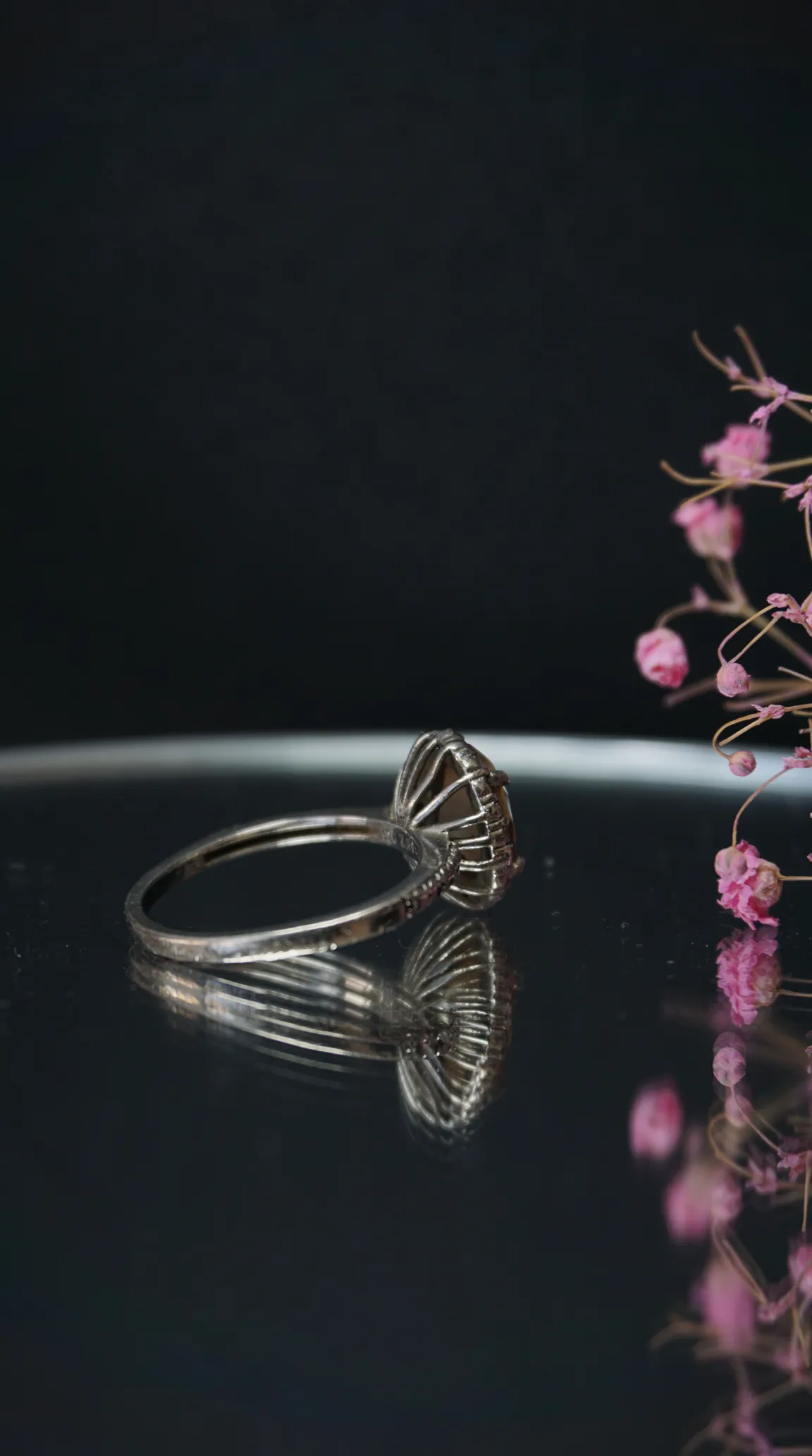 Vintage silver ring with smoky brown gemstone and intricate filigree setting, reflected on a dark surface with pink floral accents.
