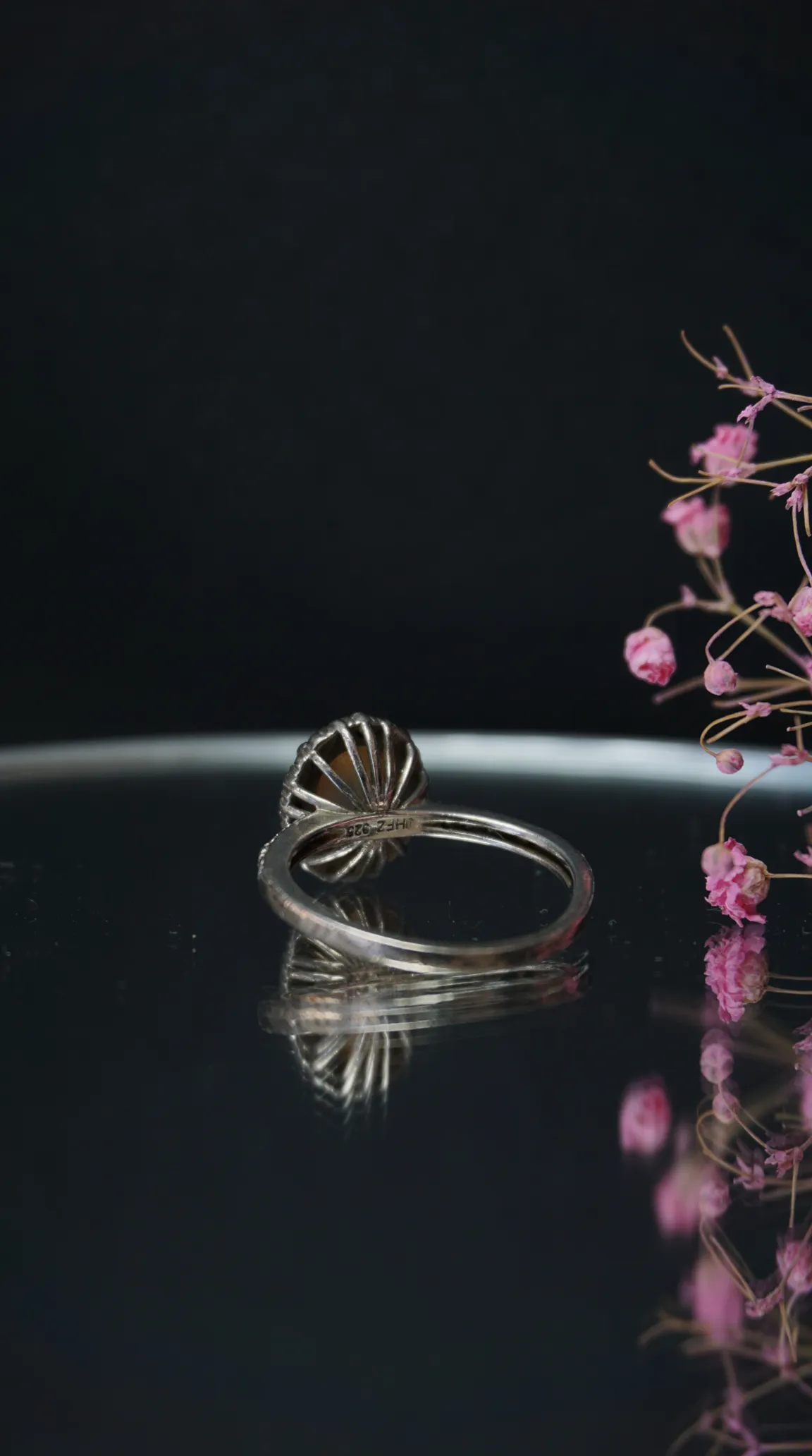 Vintage 925 sterling silver ring with a round dark brown gemstone in a fluted setting, reflecting on a glossy black surface, with dried pink flowers.