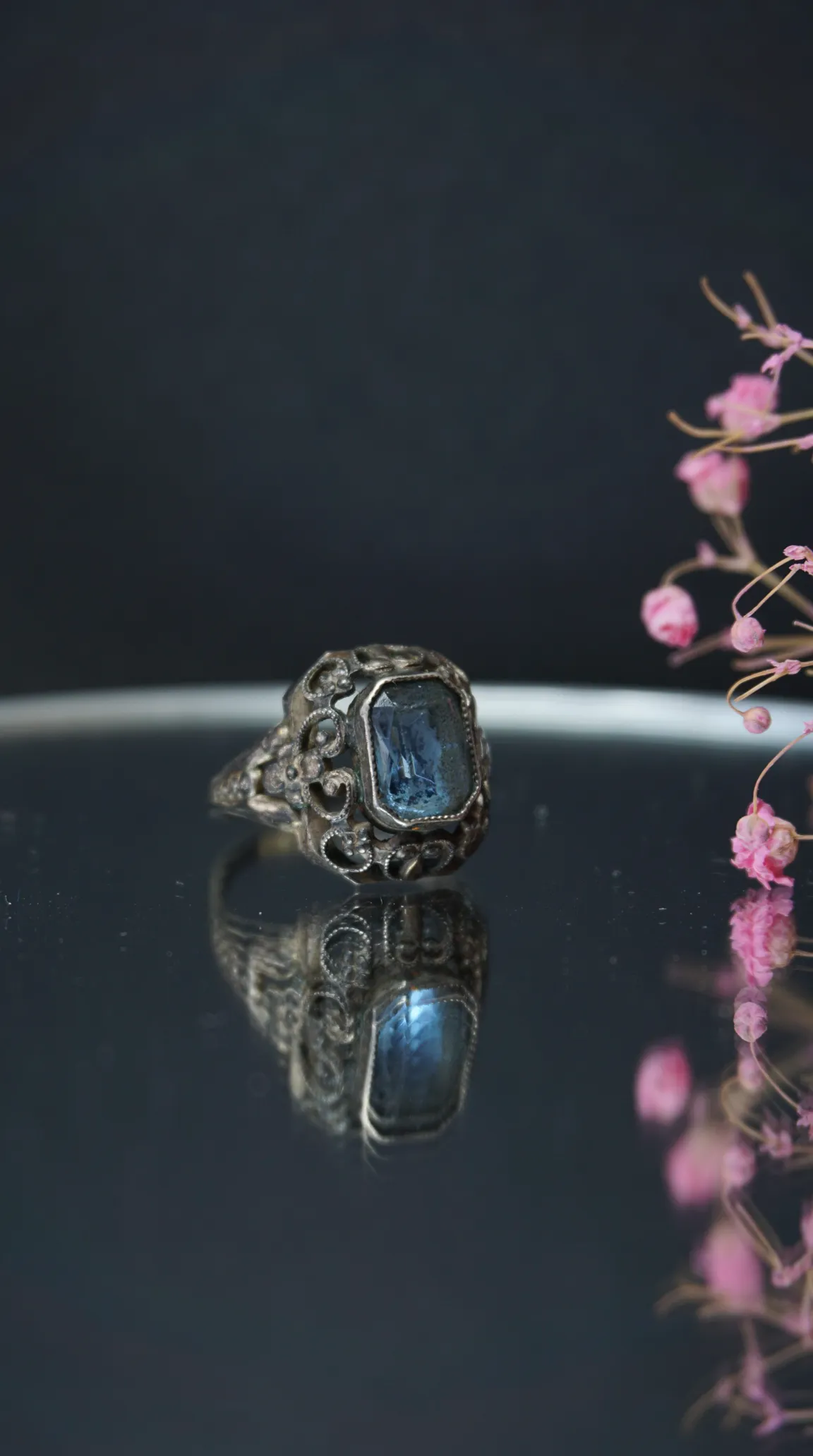 Vintage silver ring with ornate filigree and rectangular blue gemstone, reflected on dark surface with pink dried flowers.