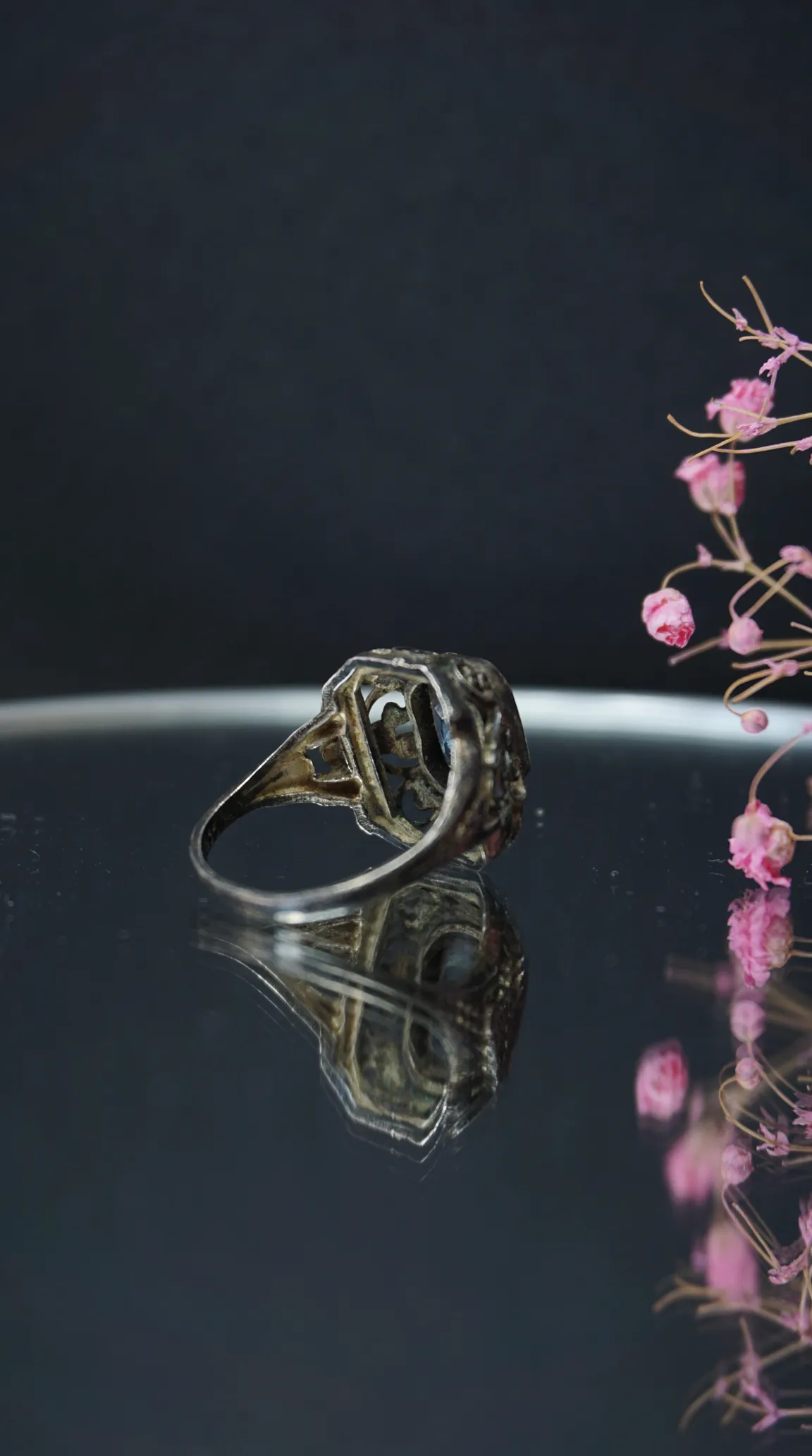 Vintage ornate filigree ring with dark gemstone and reflection, accented by pink flowers.