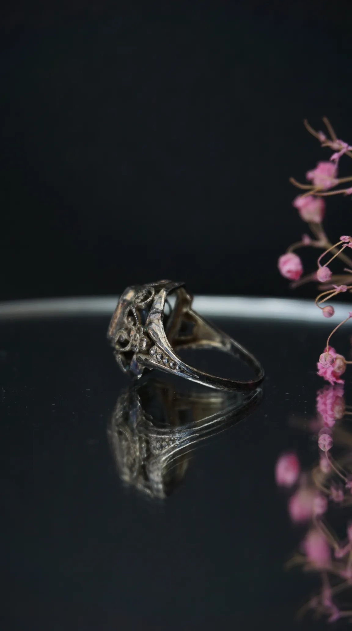 Vintage ornate silver filigree ring with a light center stone, reflected on a dark surface with blurred pink flowers.