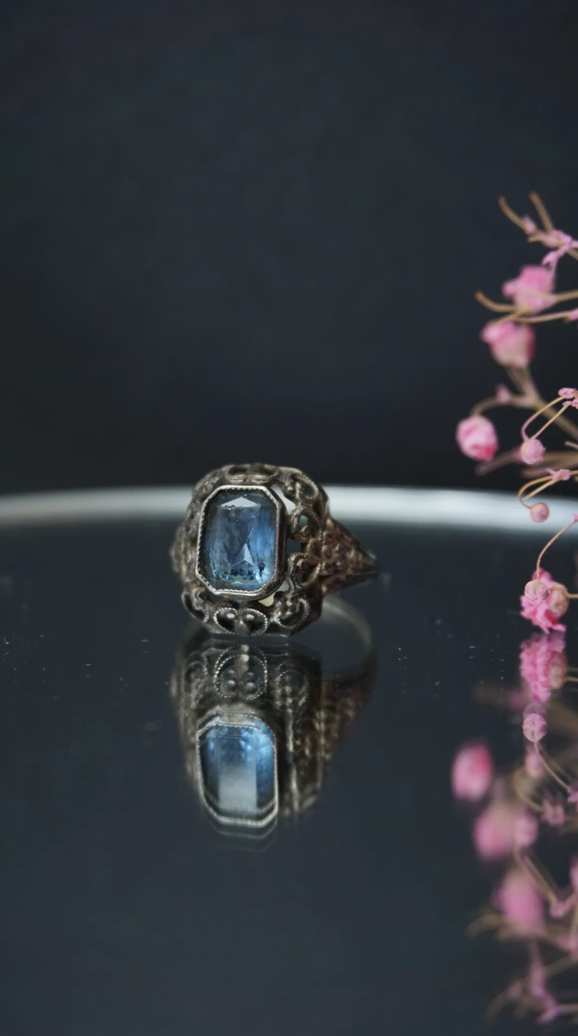 Vintage ornate silver ring with rectangular blue gemstone, reflected on dark surface beside pink flowers.