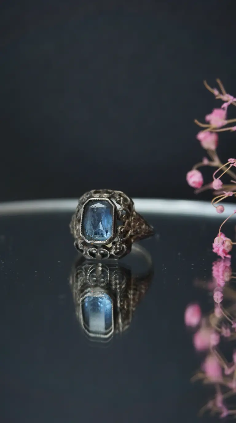 Vintage ornate silver ring with rectangular blue gemstone, reflected on dark surface beside pink flowers.