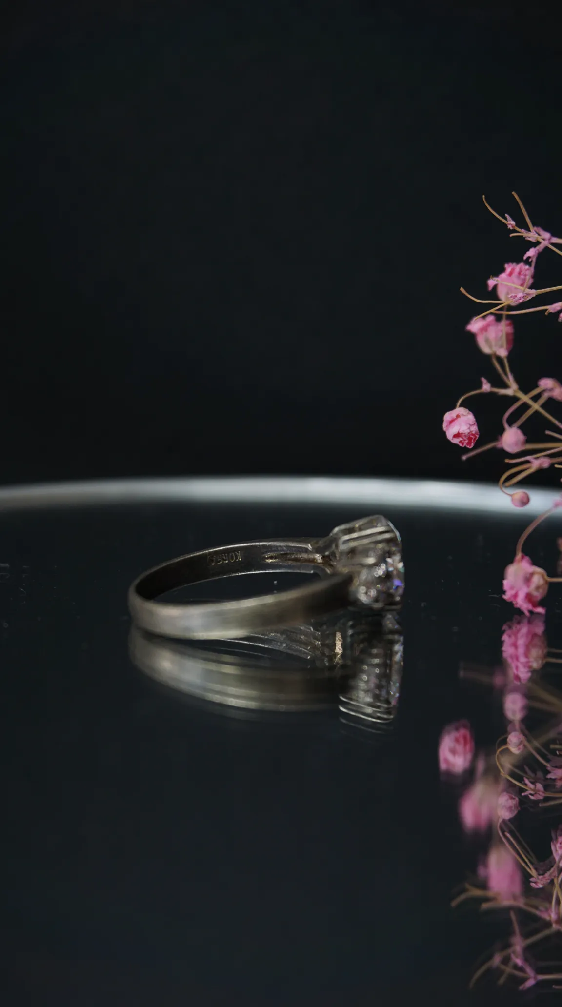 Vintage silver solitaire ring with clear gemstone, reflecting on dark surface, alongside delicate pink dried flowers. Antique jewelry.