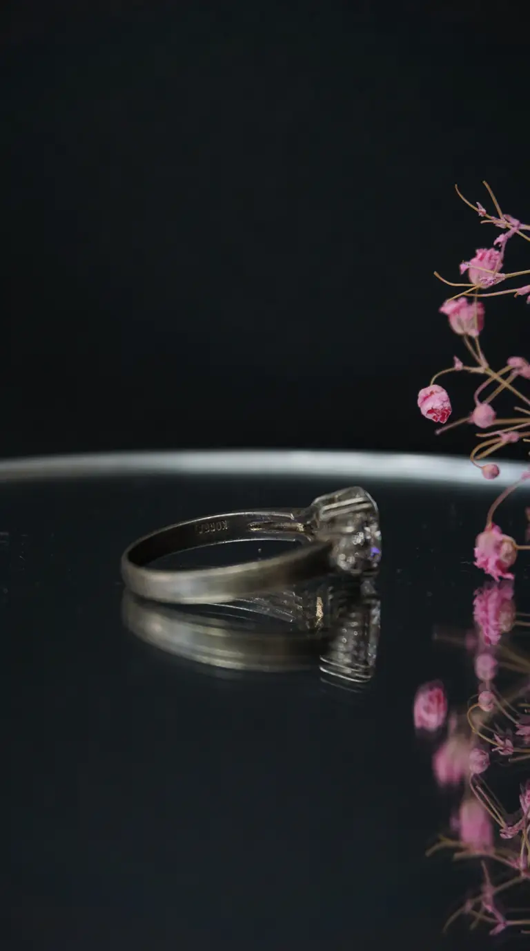 Vintage silver solitaire ring with clear gemstone, reflecting on dark surface, alongside delicate pink dried flowers. Antique jewelry.