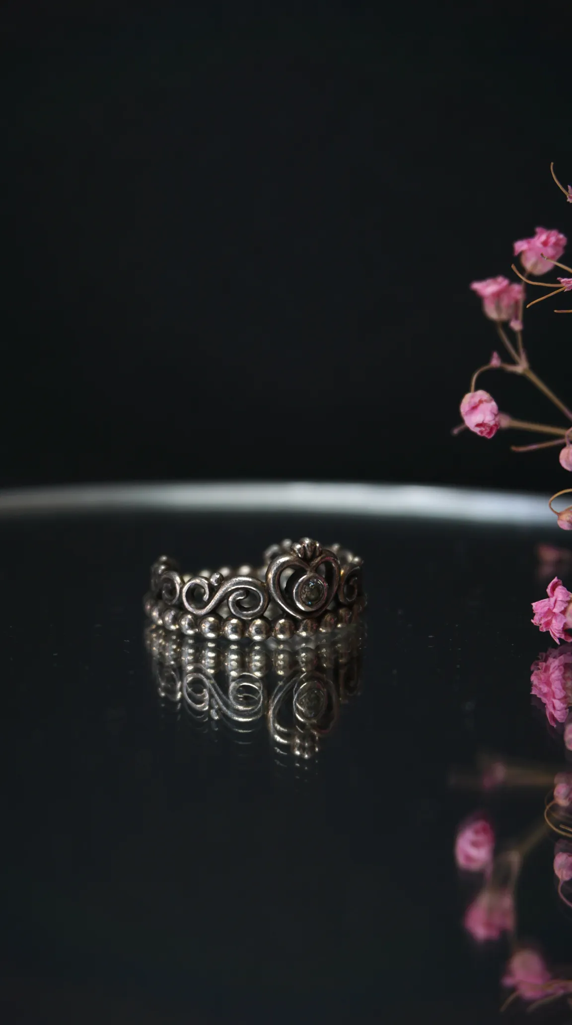 Vintage silver tiara ring with ornate scrollwork, beaded details, and a central clear gemstone, on a reflective dark surface with soft pink dried flowers.