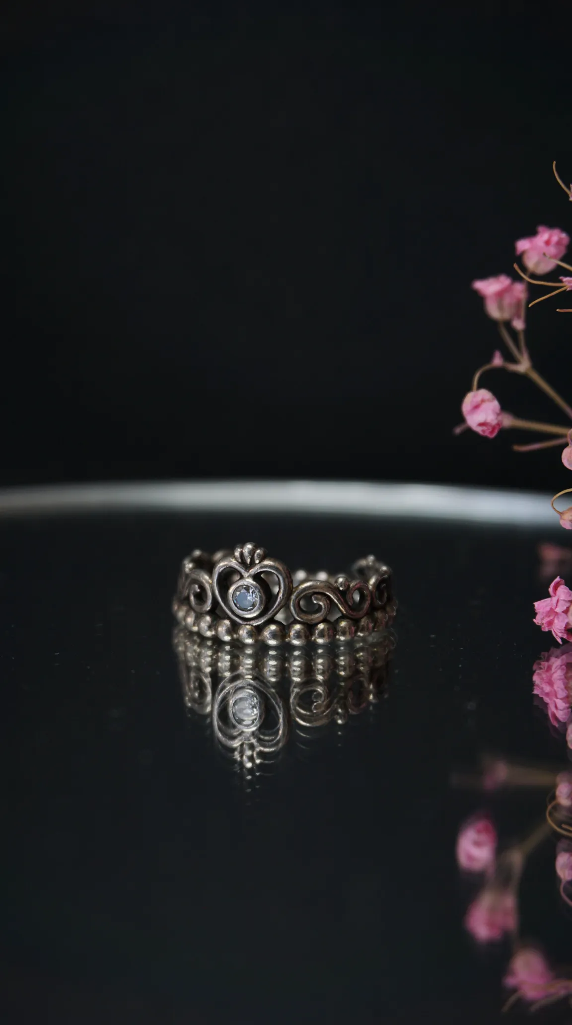 Vintage sterling silver crown ring with heart and swirl design, featuring a clear gemstone, reflected on a dark surface.
