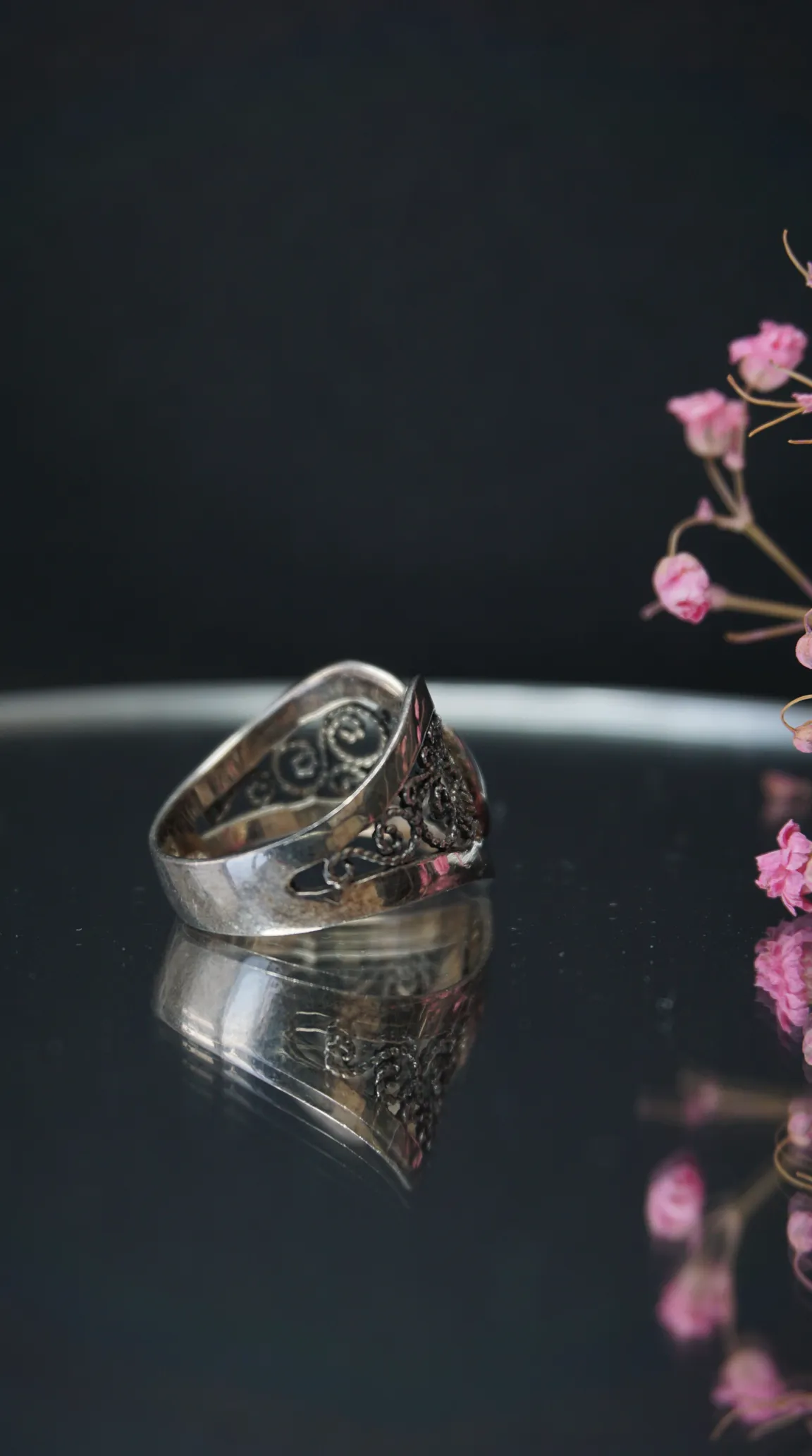 Vintage silver filigree ring with ornate scrollwork, reflected on dark surface, with pink dried flowers.