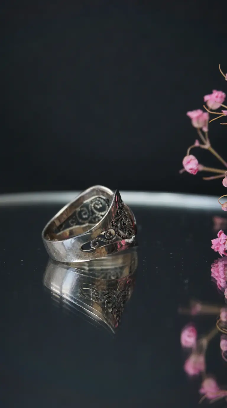 Vintage silver filigree ring with ornate scrollwork, reflected on dark surface, with pink dried flowers.