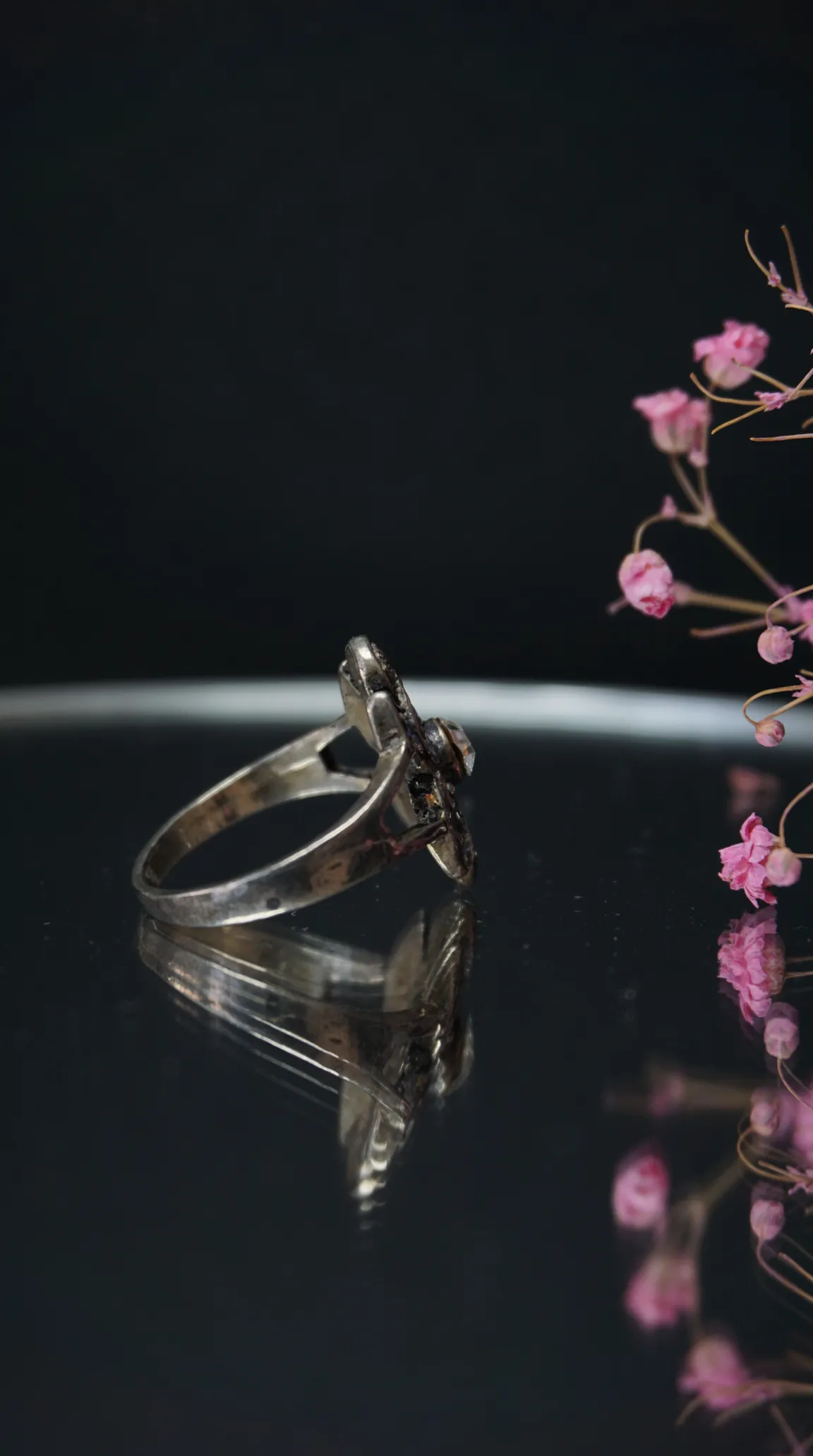 Vintage silver ring with ornate design and dark stone, reflecting on a black surface beside pink dried flowers.