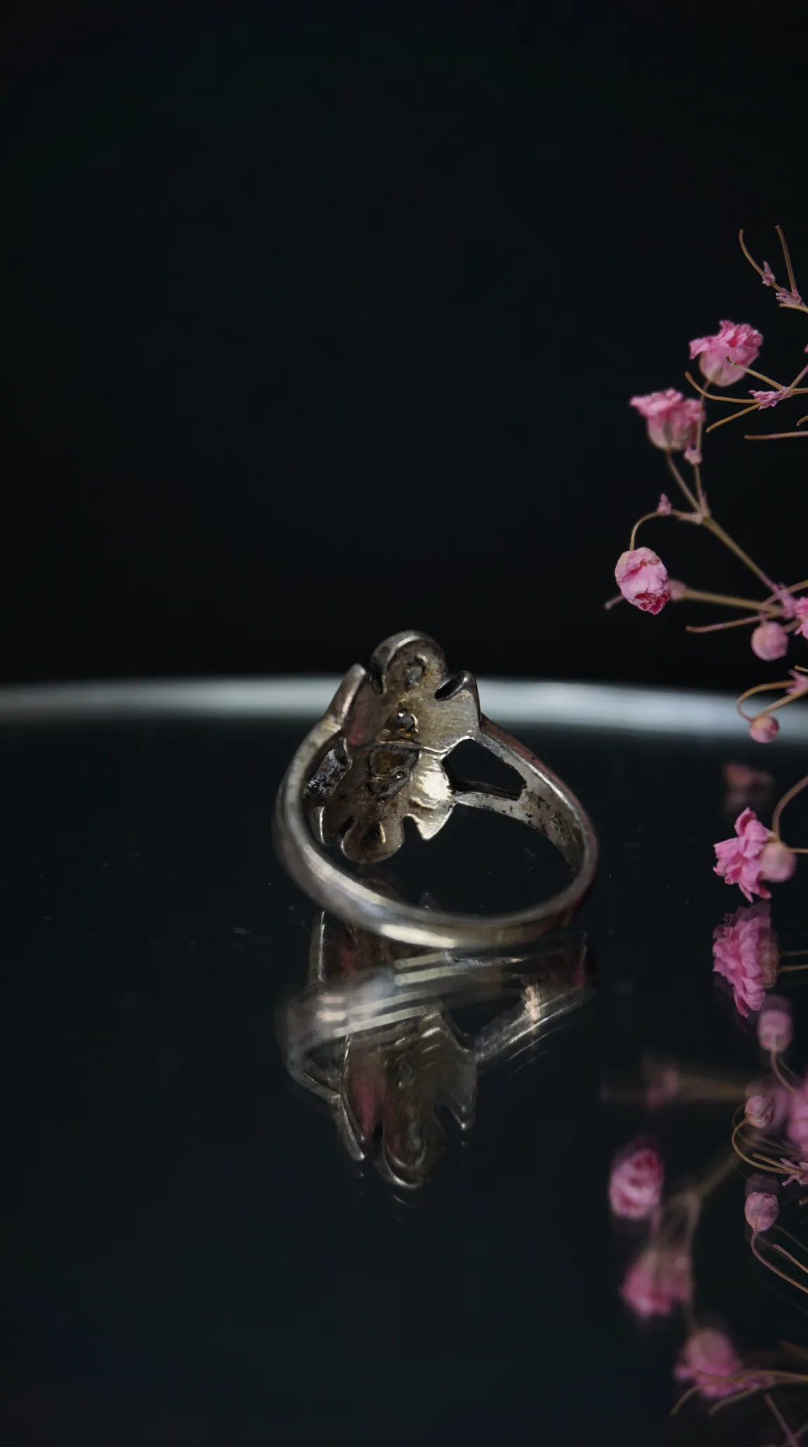 Vintage silver ornate ring on reflective black surface with pink dried flowers.