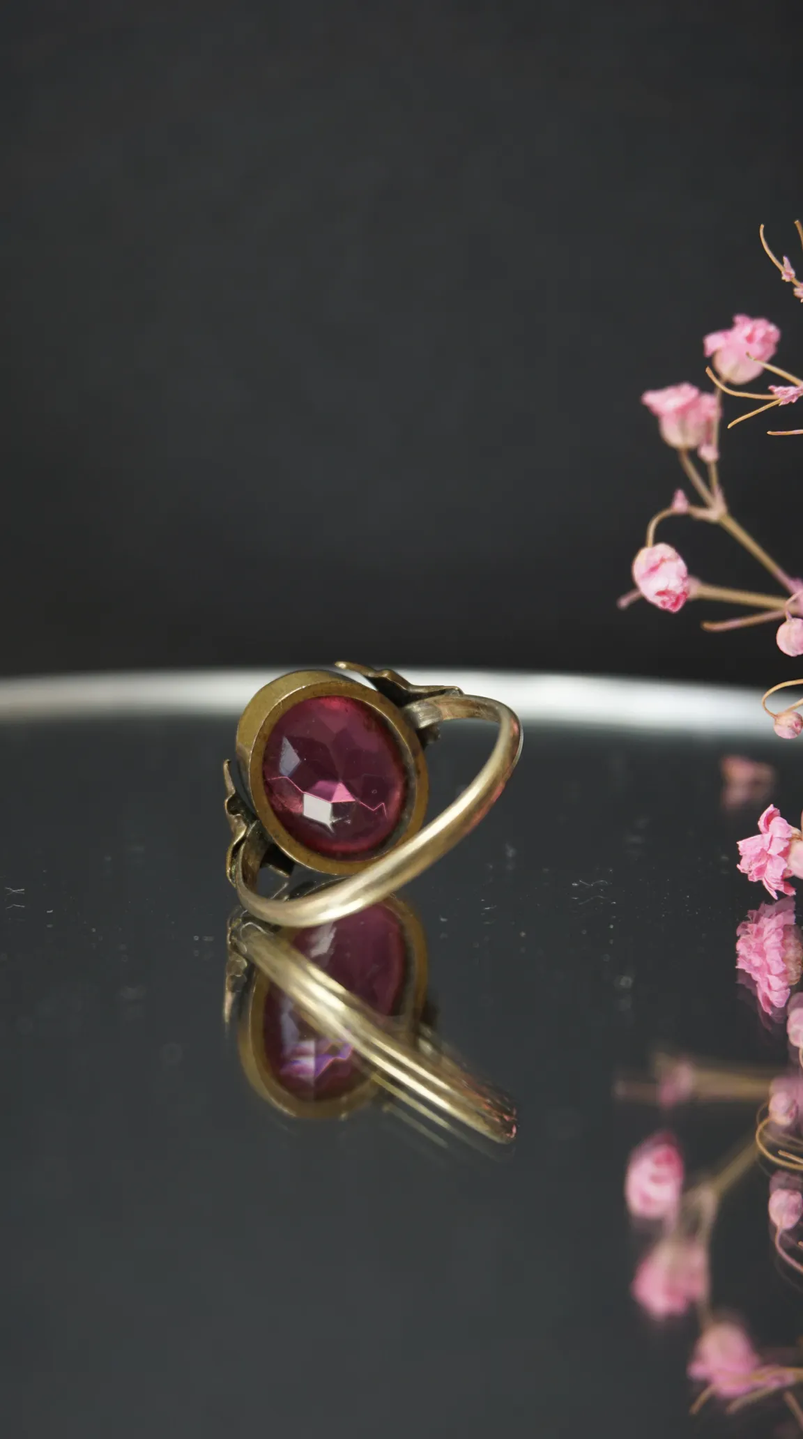 Vintage gold tone ring with large faceted pink gemstone, reflecting on dark surface, with dried pink flowers. Antique jewelry.