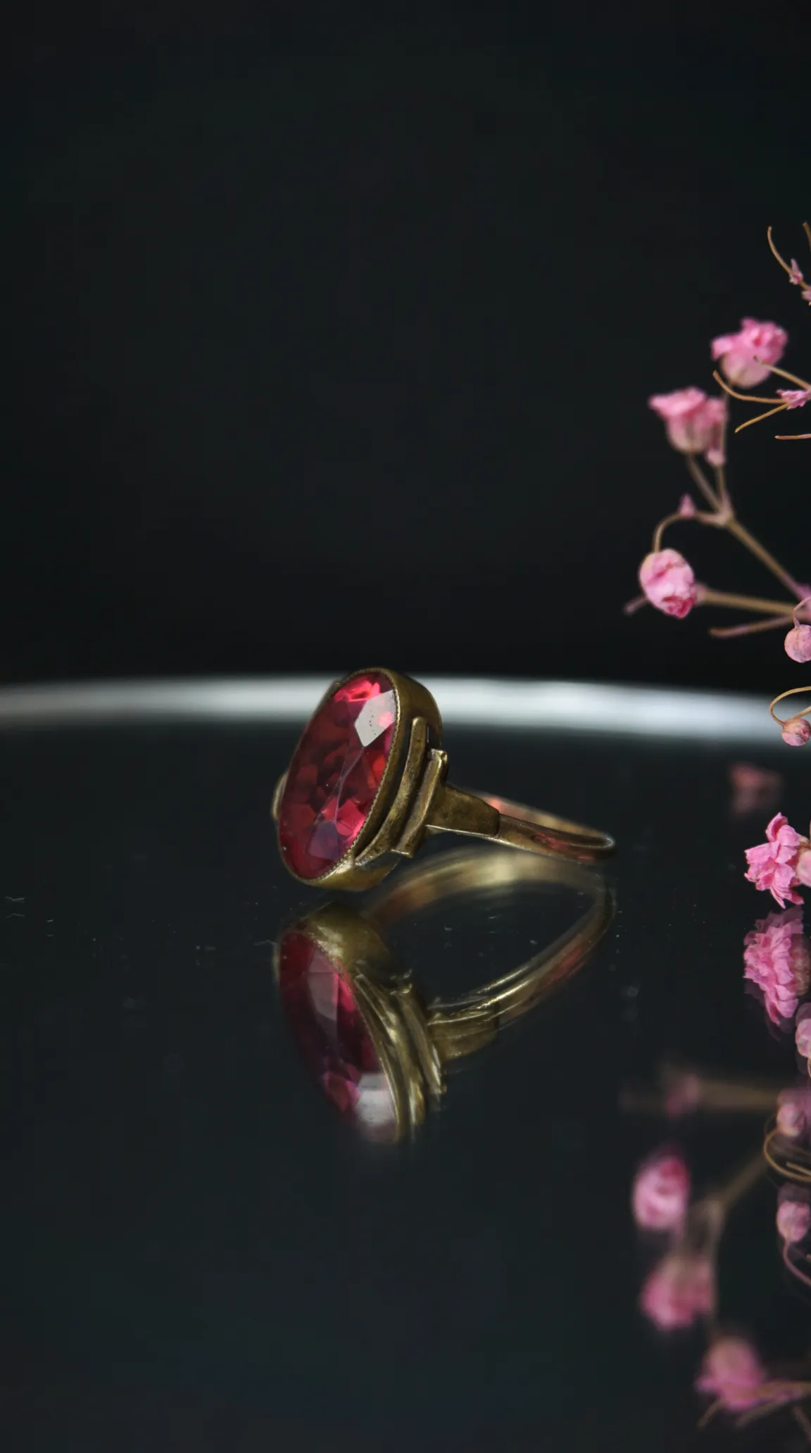 Vintage gold tone ring with large oval red gemstone, possibly ruby or garnet, on a dark reflective surface.