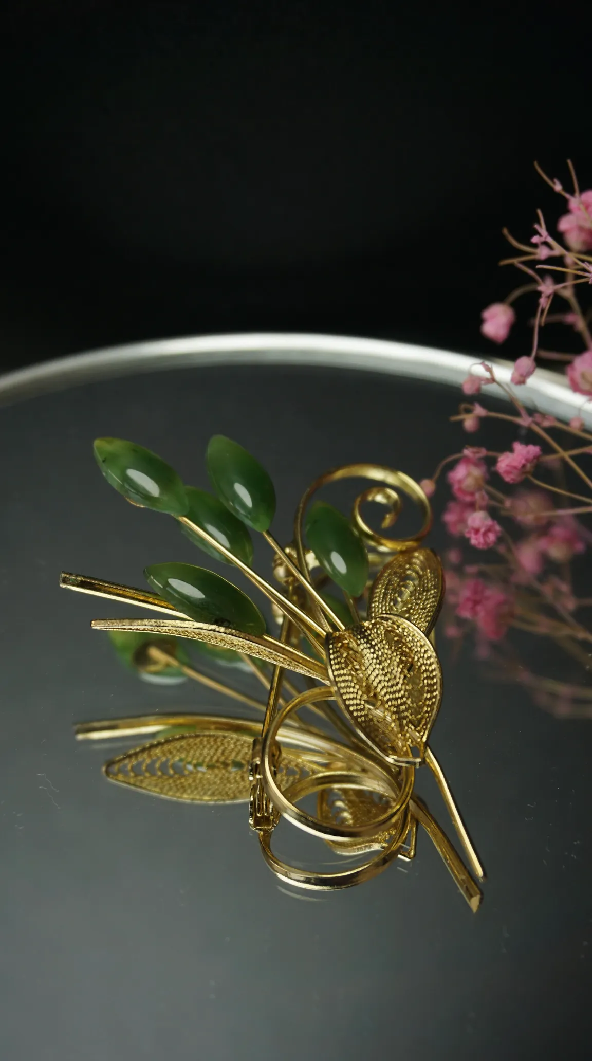Vintage gold-tone floral brooch with green jade leaves and filigree details, on a reflective surface.