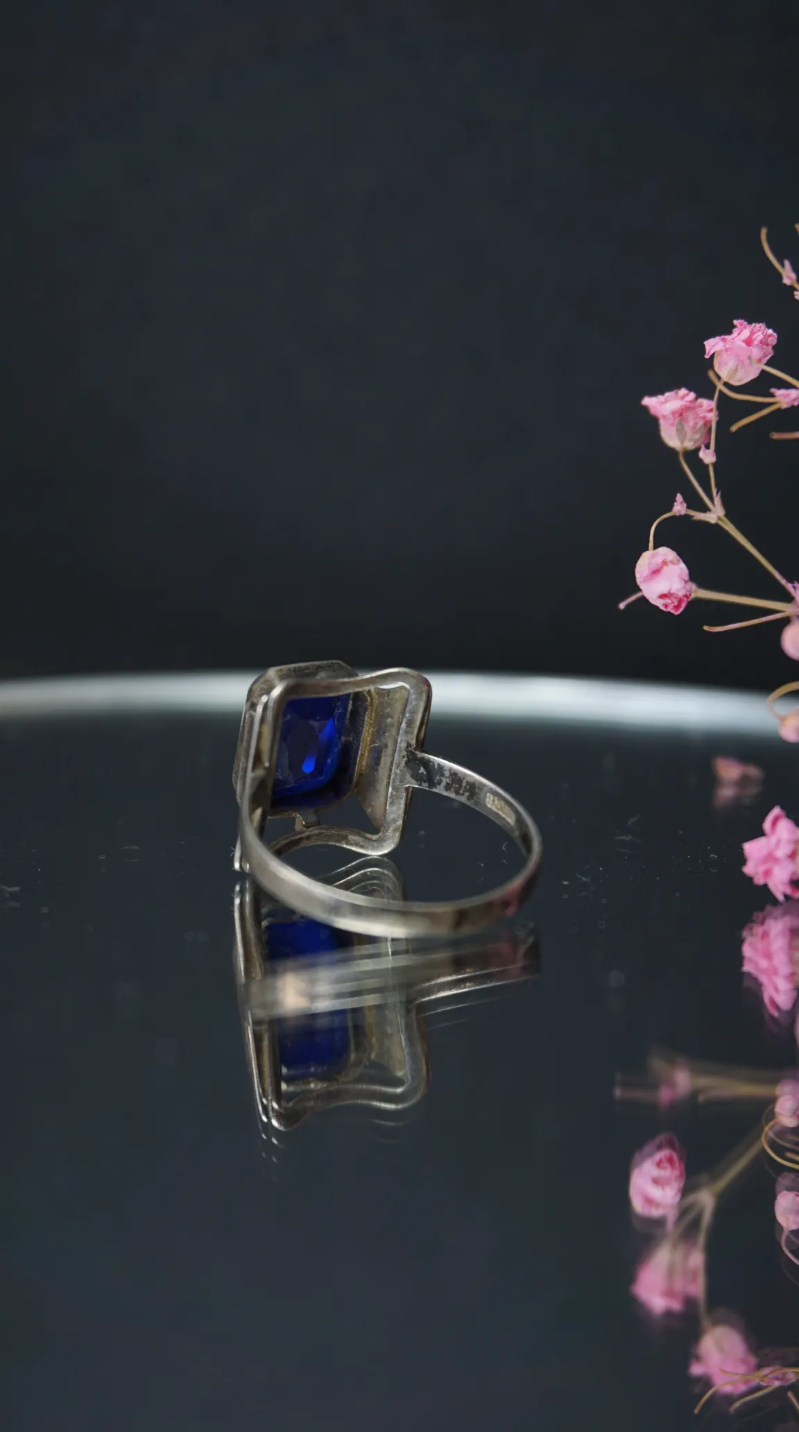 Vintage sterling silver ring with rectangular deep blue sapphire gemstone, reflecting on a polished surface, styled with dried pink flowers.