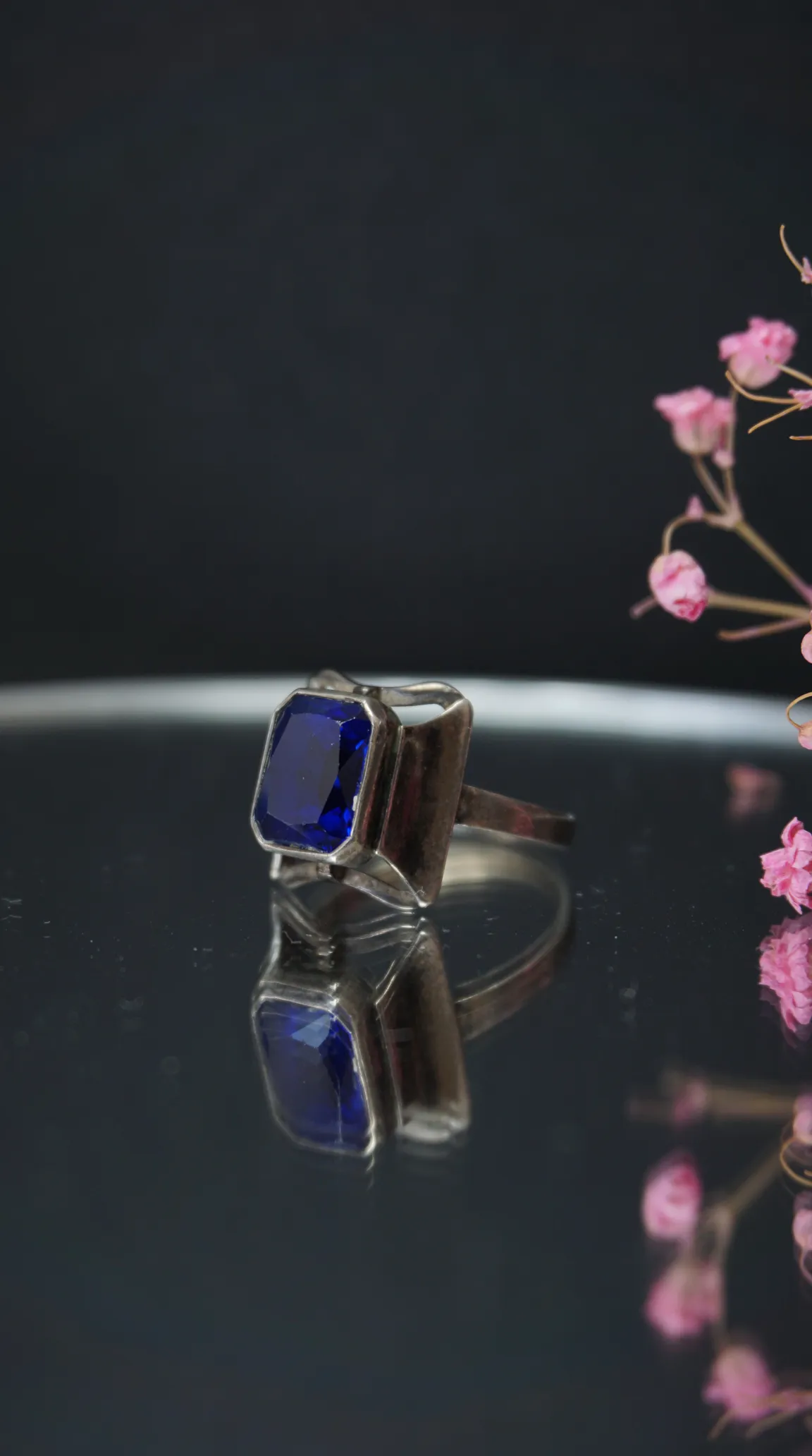 Vintage sterling silver ring with rectangular royal blue sapphire gemstone, reflecting on a dark surface with pink flowers in background.