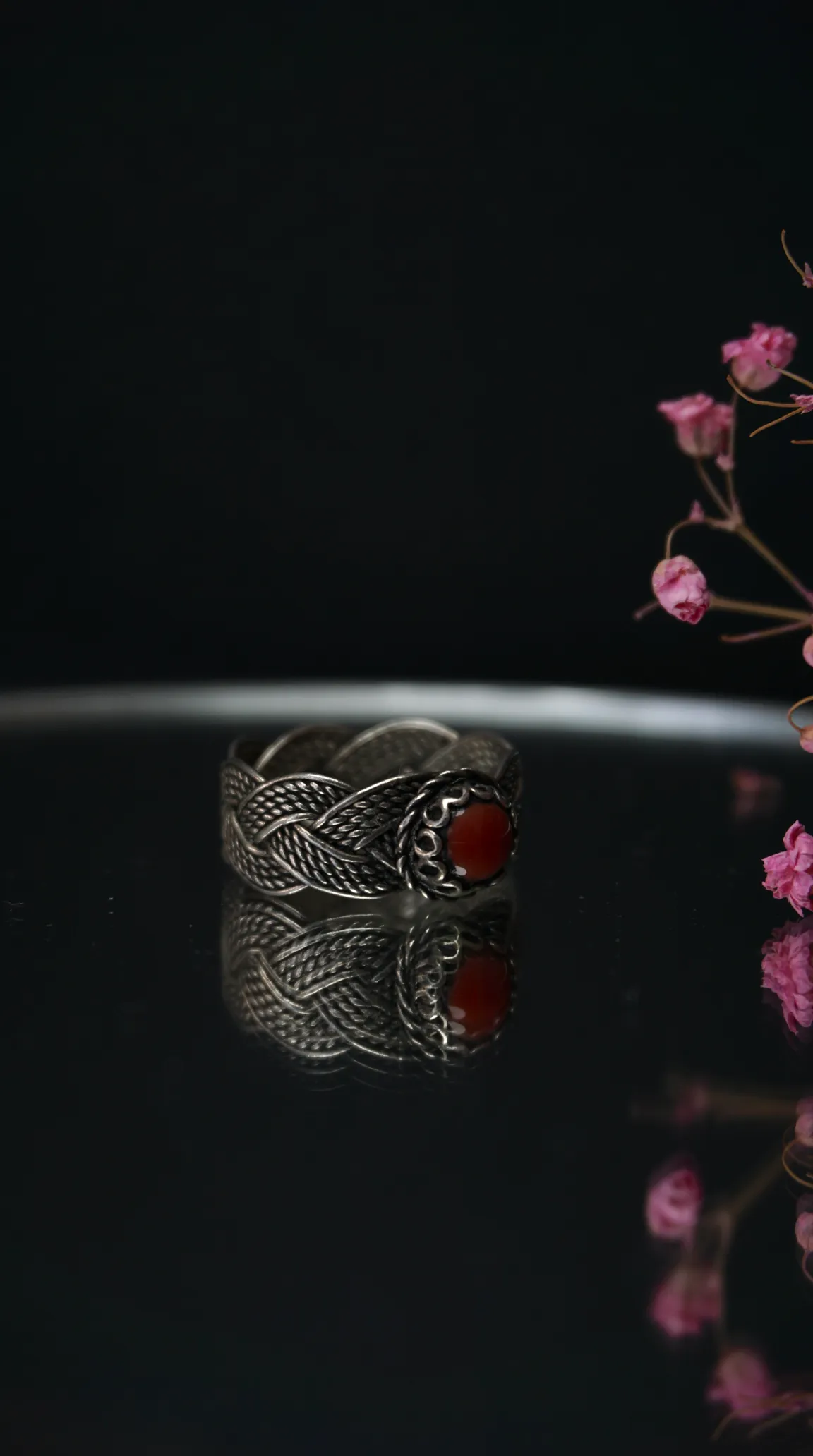 Vintage braided silver ring with red cabochon gemstone on dark reflective surface.