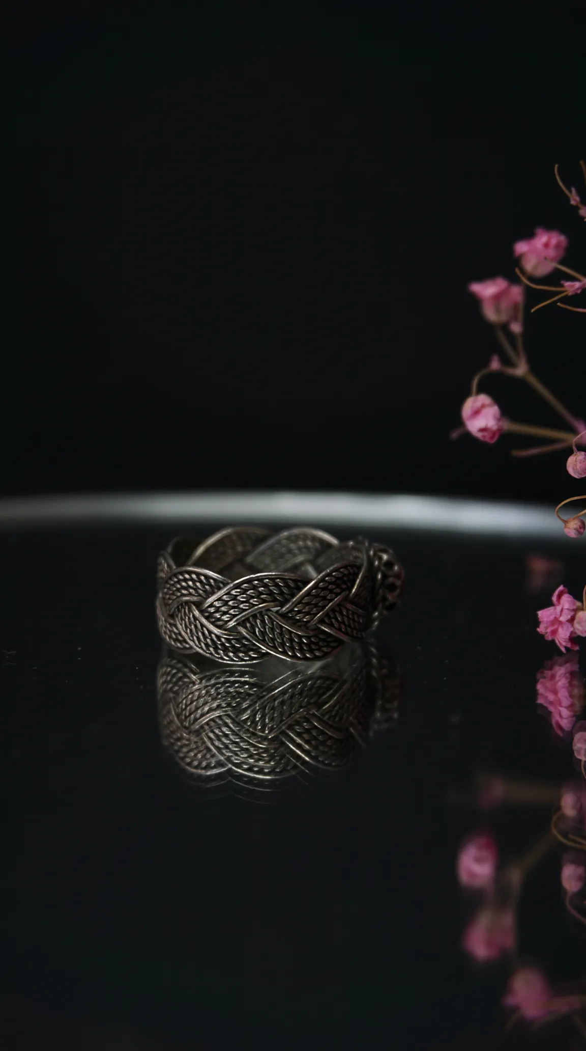 Vintage silver braided ring with intricate woven design, reflecting on a dark surface, with delicate pink flowers.